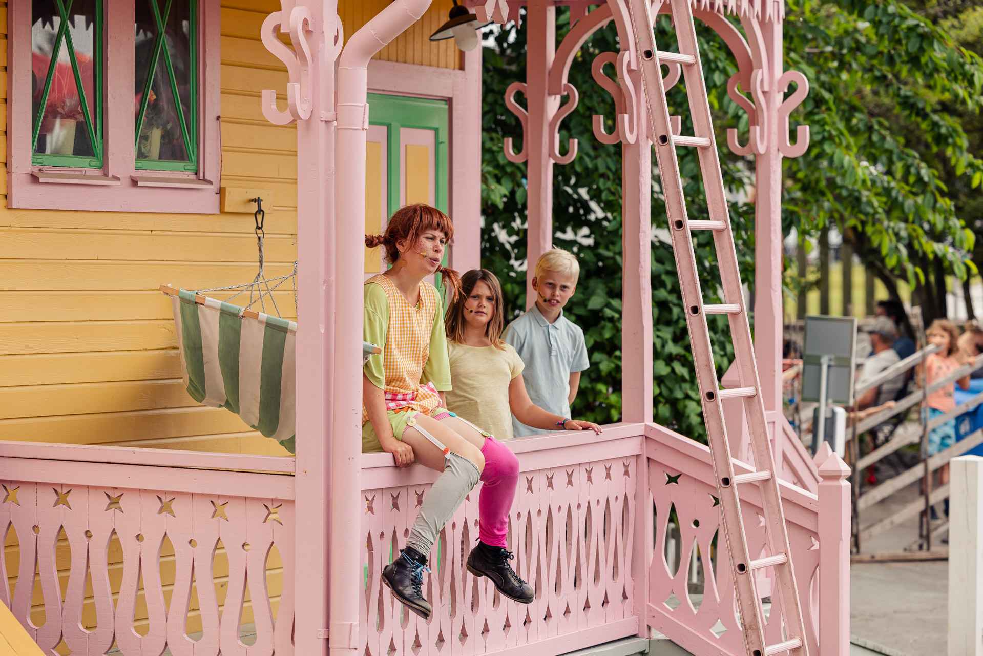 Een theatervoorstelling met Pippi Langkous die op de veranda van haar gele huis zit, Tommy en Annika staan naast haar.