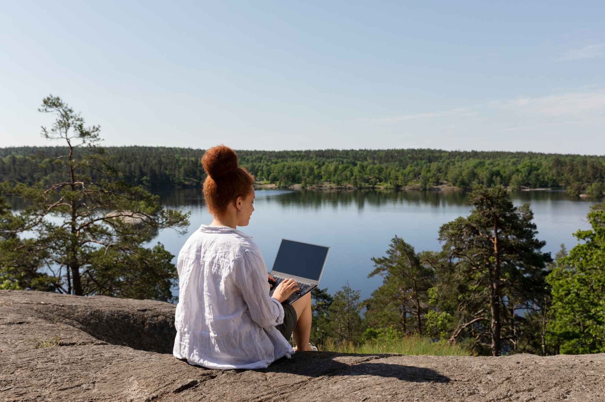 Een vrouw zit op een rots met haar laptop op haar schoot. Op de achtergrond is een meer met bos rond.