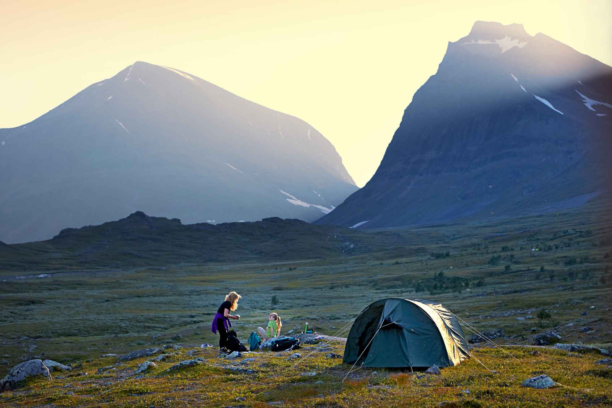Een tent staat midden in de natuur. Op de achtergrond twee bergen. Twee personen zitten naast de tent.