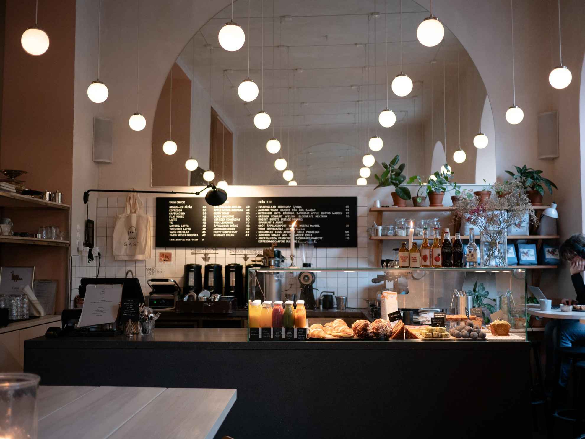 Een toonbank met broodjes, brood, gebak en diverse frisdranken in een café. Achter de toonbank een koffiezetapparaat. Aan het plafond hangen verschillende kleine bollampjes.
