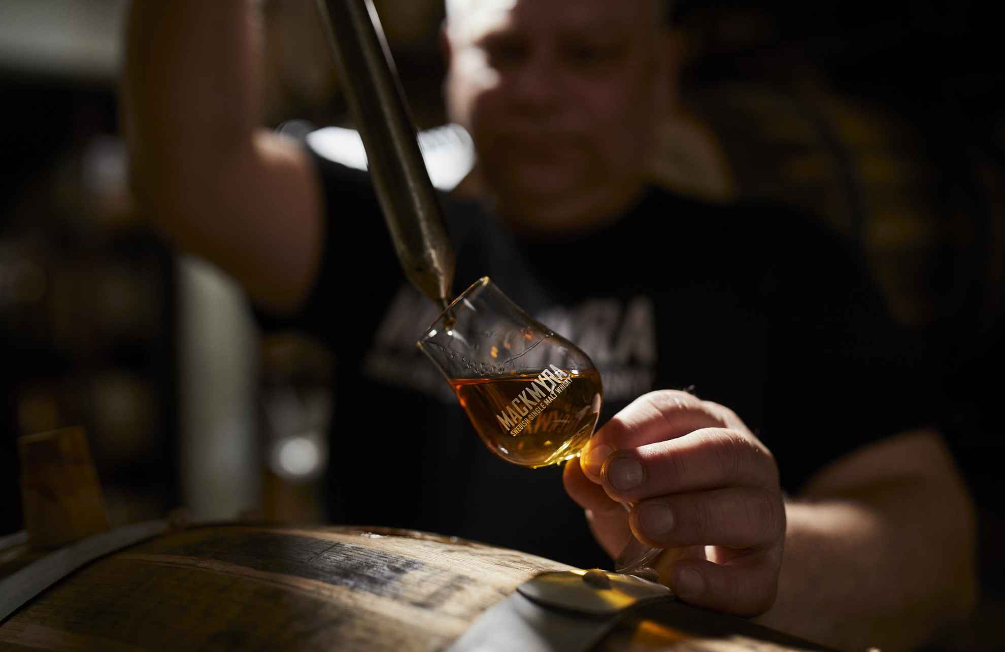 Whisky wordt uit een pipet in een whiskyglas gegoten met de tekst Mackmyra erop. Op de voorgrond staat een eiken vat.