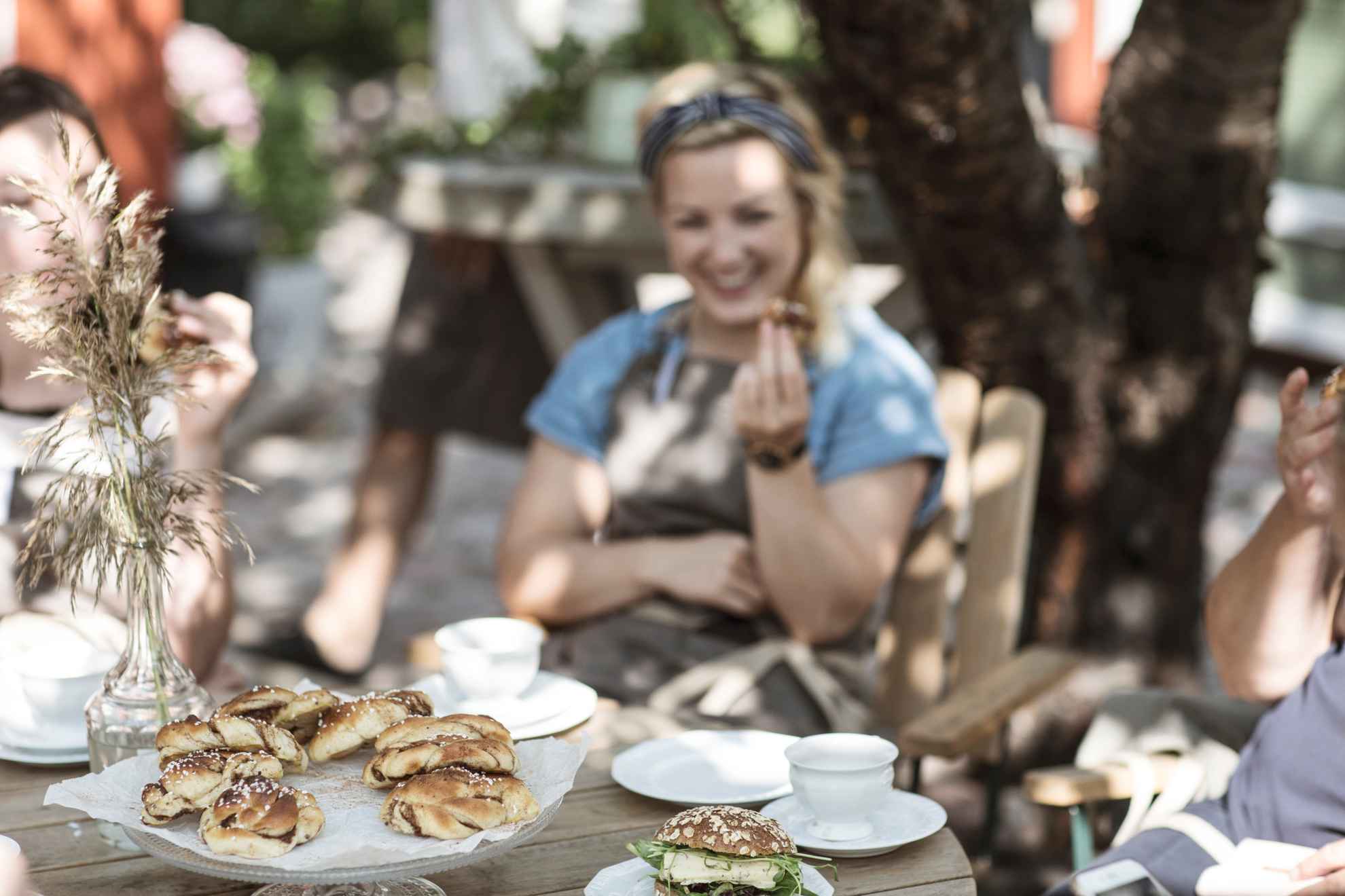 Een vrouw zit aan een tafel buiten een kaneelbroodje te eten. Op de tafels staan een bord met kaneelbroodjes, een broodje en twee koffiekopjes.