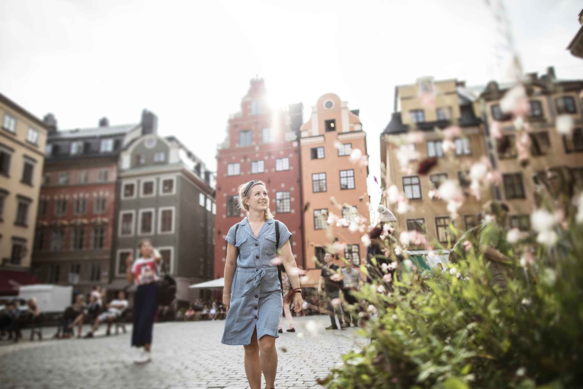 Een vrouw loopt op een plein in de oude stad. Op de achtergrond zie je oude stenen huizen en vooraan staat een bloeiende plant.