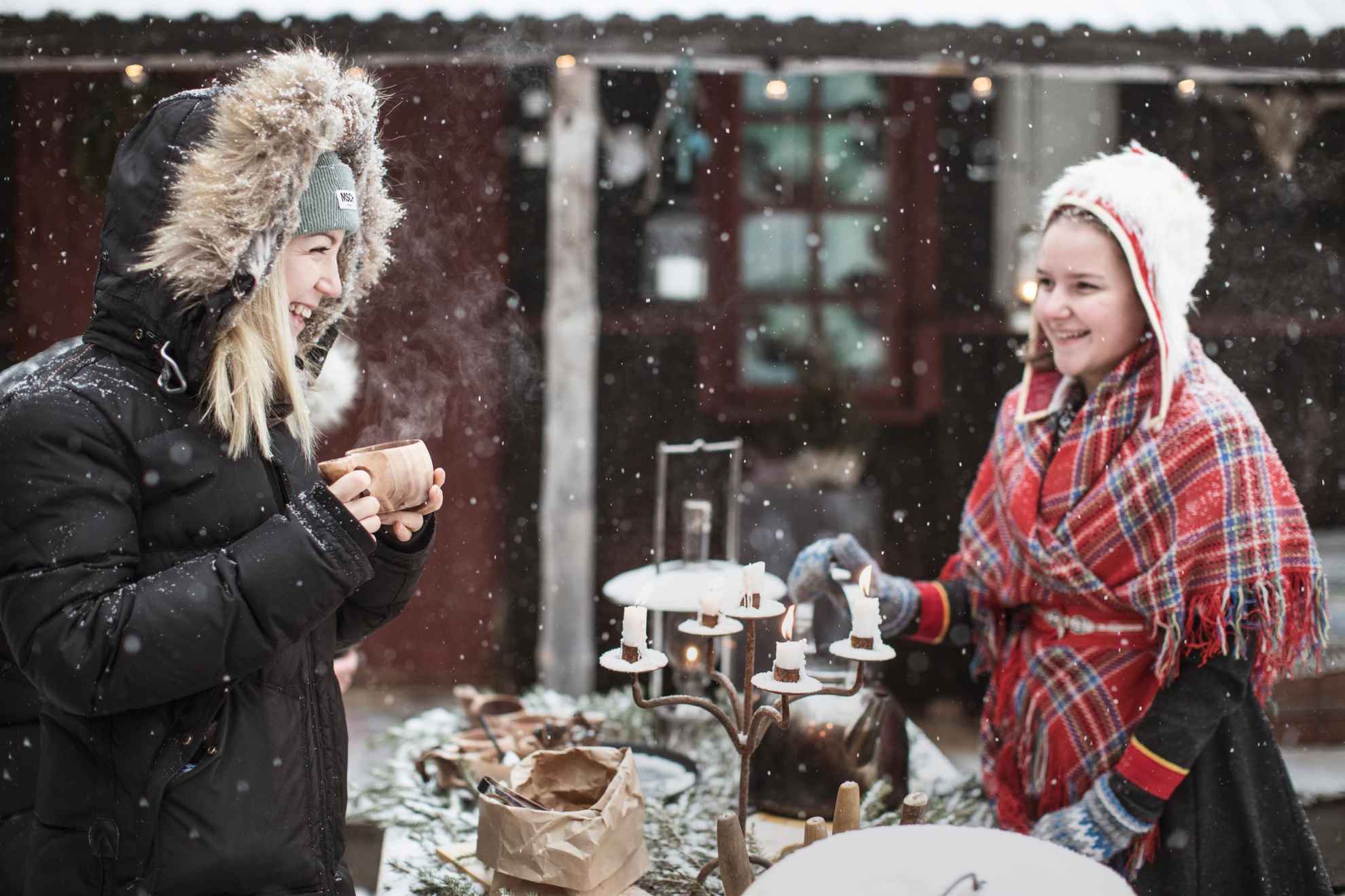 Twee vrouwen staan aan een tafel met een fika buiten in de sneeuw. Op tafel staat een oude kandelaar met brandende kaarsen. Een van de meisjes draagt een Sámi-kostuum. Het sneeuwt.