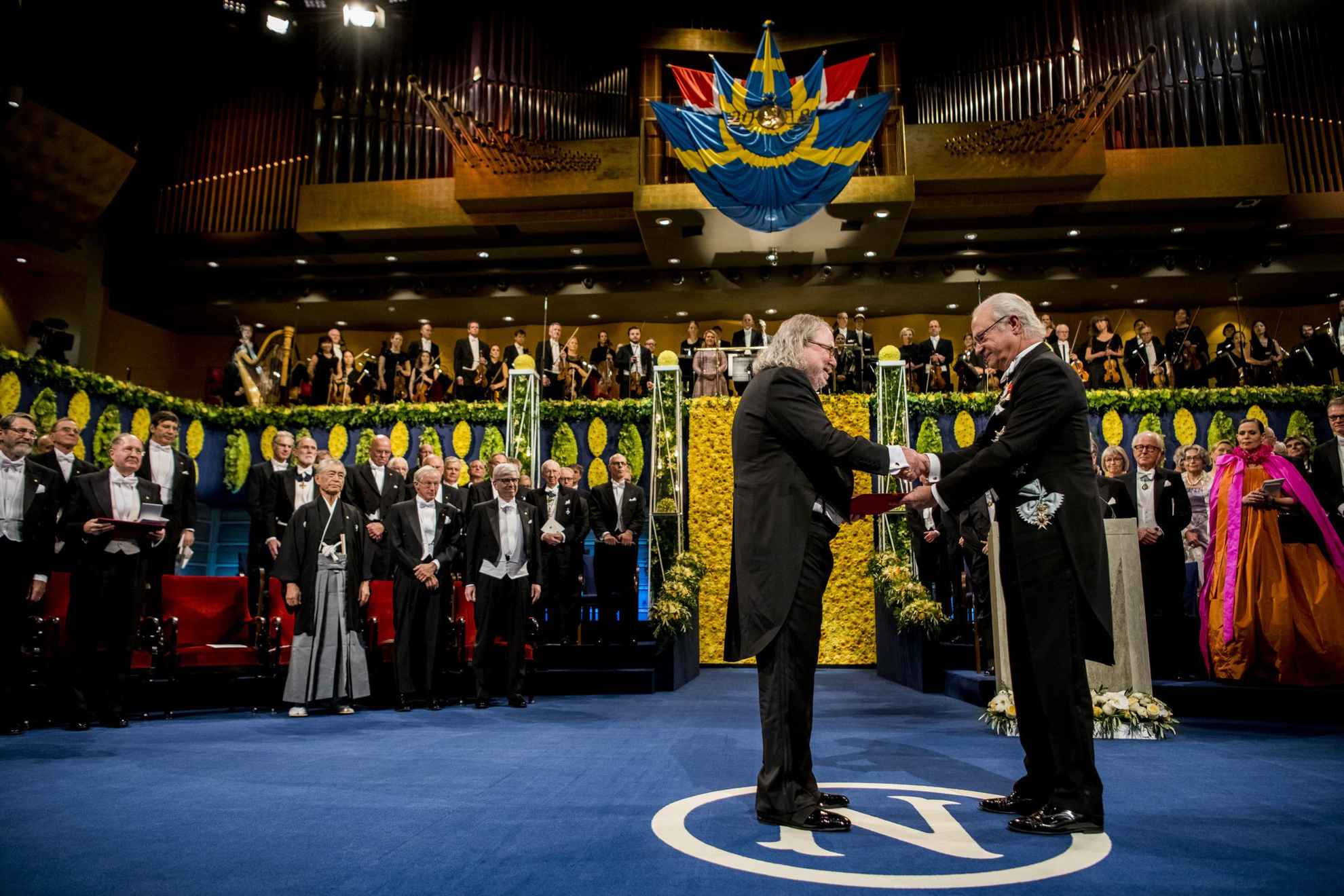 Een mannelijke laureaat staat op een podium en ontvangt zijn prijs uit handen van de Zweedse koning Carl XVI Gustaf.