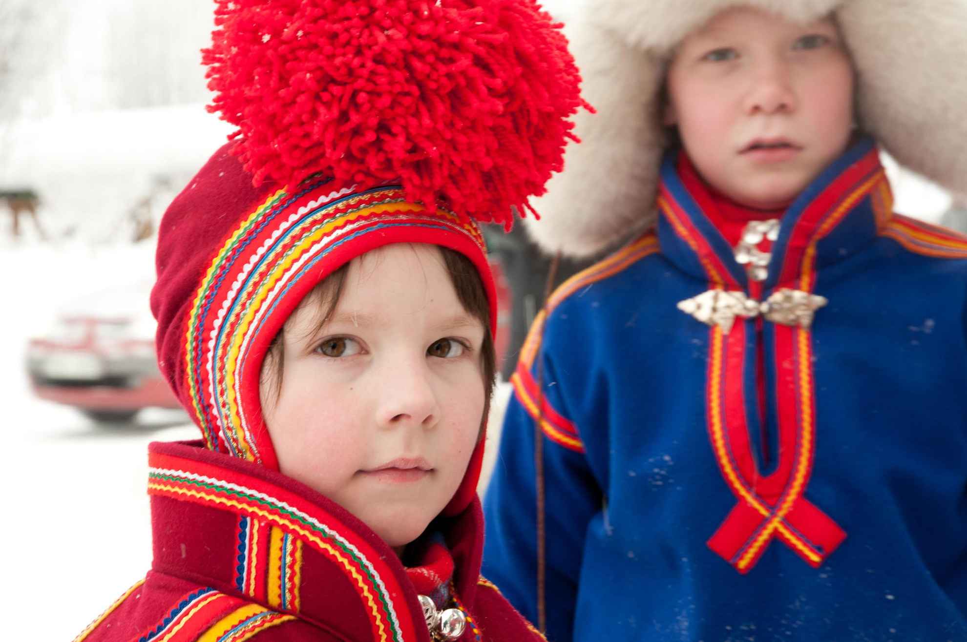 Close-up van een Sami-kind in hun rode, traditionele klederdracht. Wazig op de achtergrond nog een kind met een blauw klederdracht.