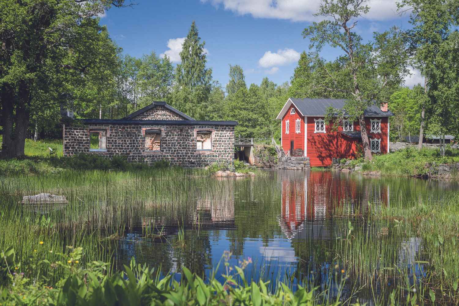 Een rood houten huis met witte versieringen en een oud stenen huis naast een meer in de zomer.