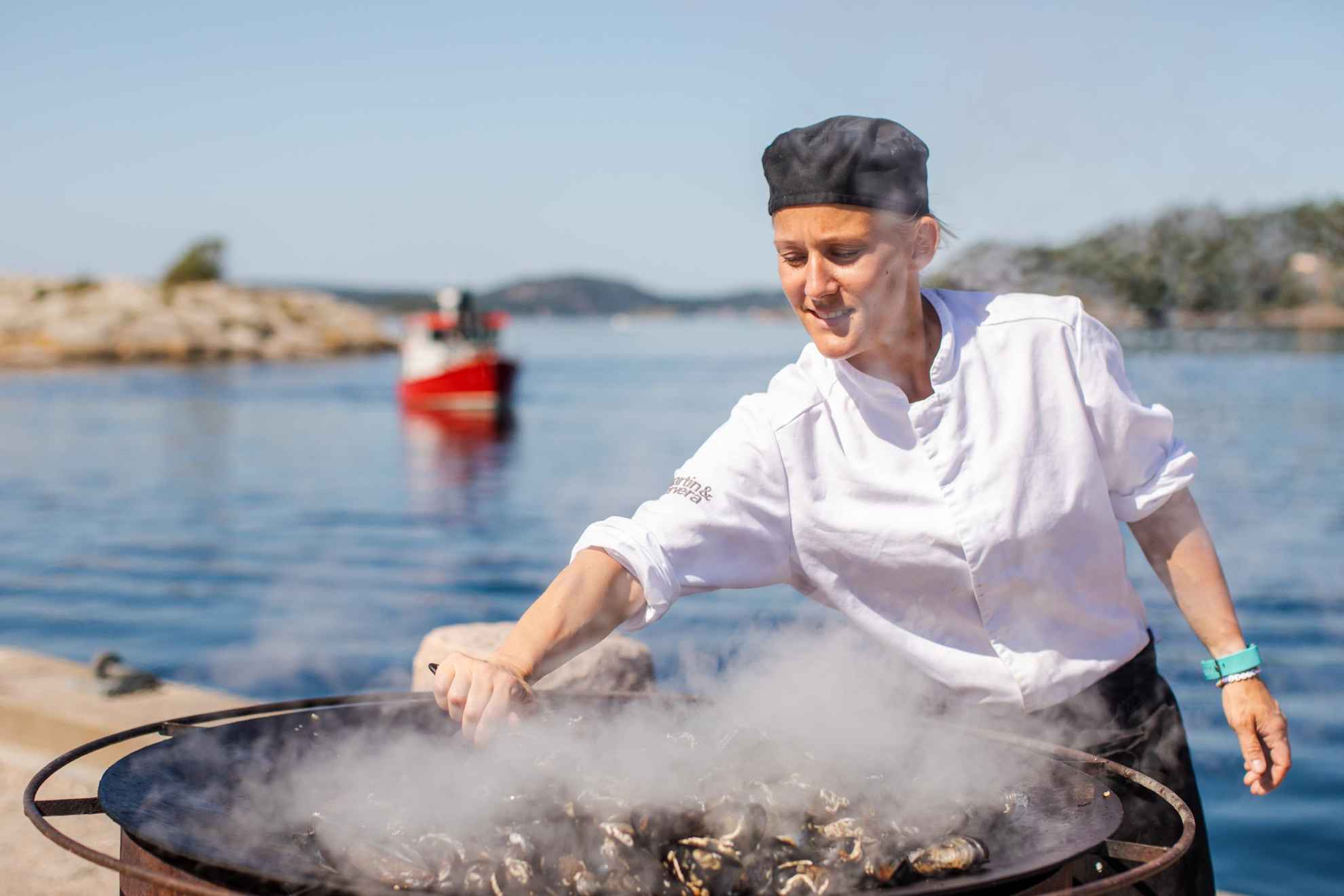 Een chef-kok staat buiten bij de zee mosselen te koken in een grote pan.