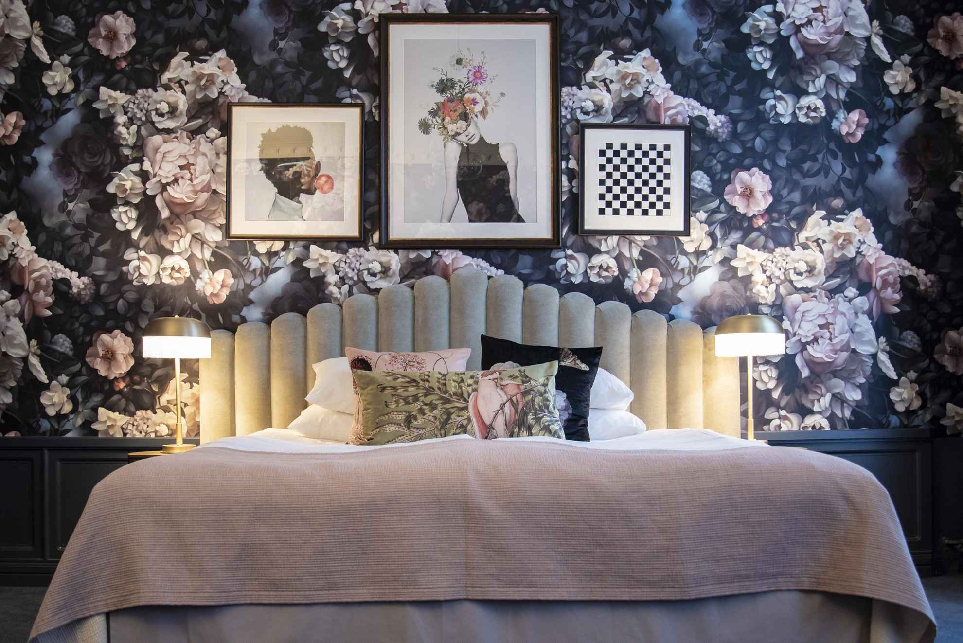 Het interieur van een kamer in een hotel. Een bed voor een muur met een behang met grote roze en witte rozen. Er hangen drie foto's aan de muur en drie kussens met verschillende patronen op het bed.
