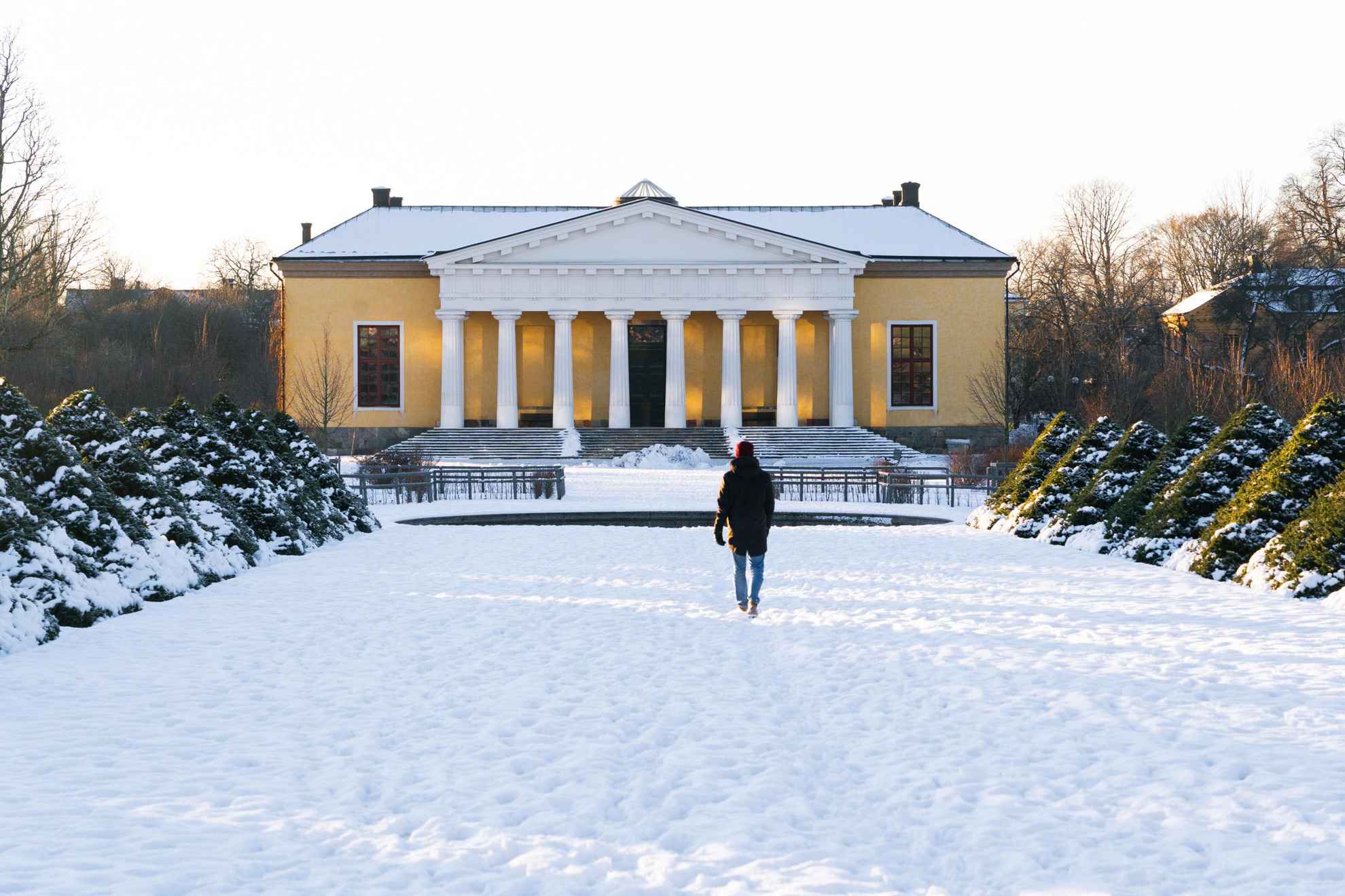 Een persoon loopt in de winter naar een geel huis in de botanische tuin van de Universiteit van Uppsala.