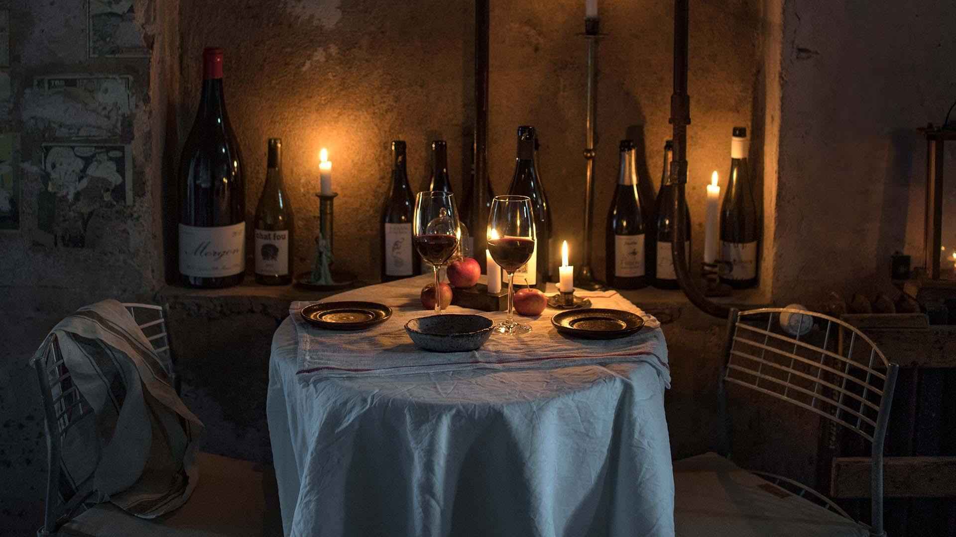 Een tafel voor twee met een wit tafelkleed, borden en twee glazen gevuld met rode wijn. De muur is versierd met lege wijnflessen en brandende kaarsen.