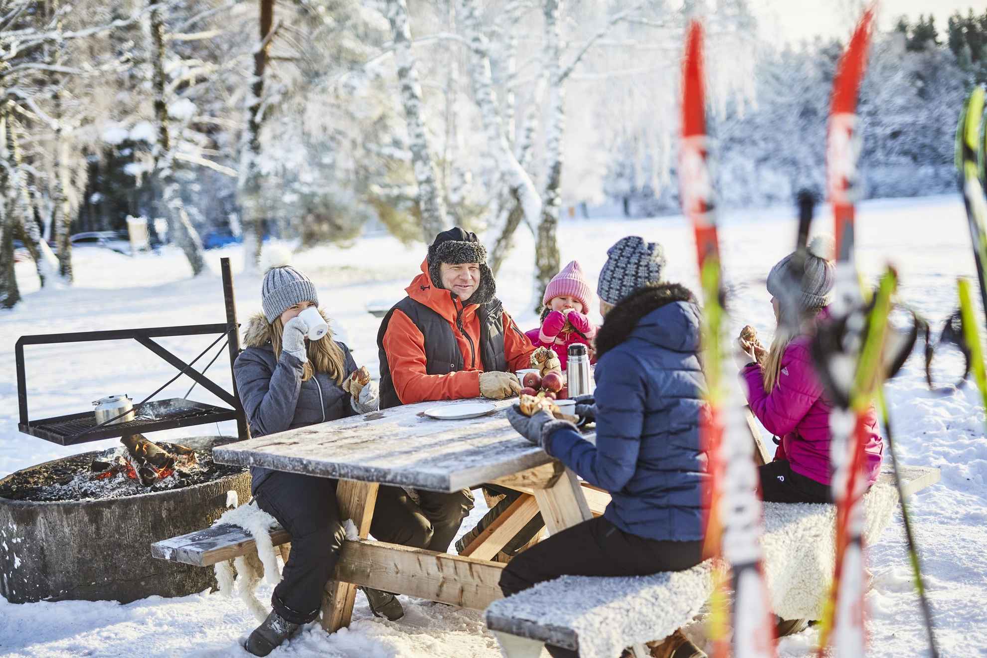 Een gezin van vijf zit buiten aan een tafel, kaneelbroodjes eten en koffie drinken. Er zijn ski's op de voorgrond van de foto.