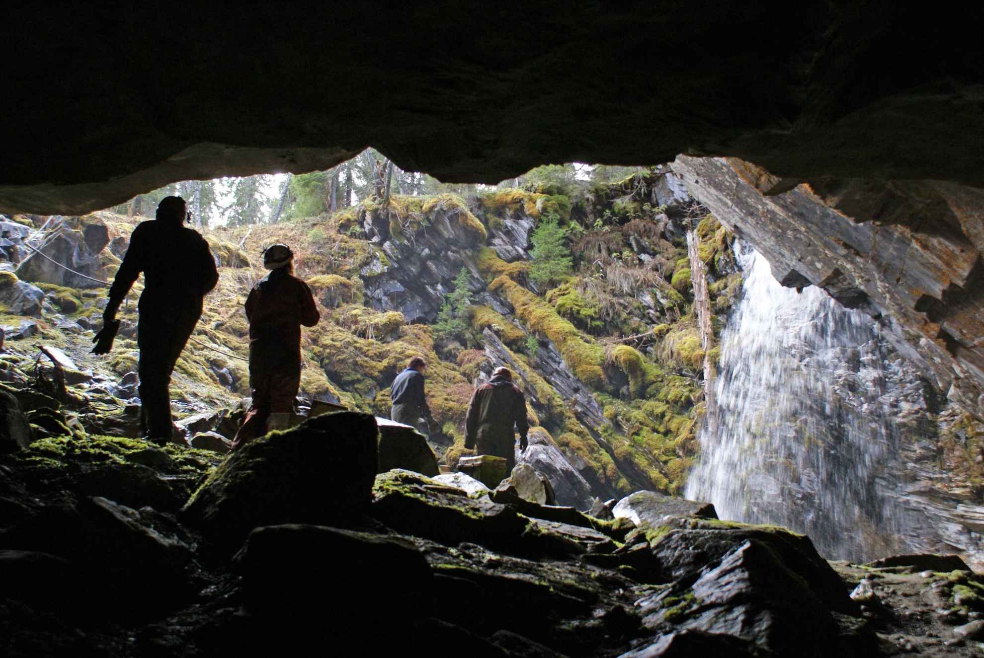 Een blik vanuit een grot naar een open plek met vier mensen die naar een waterval lopen.