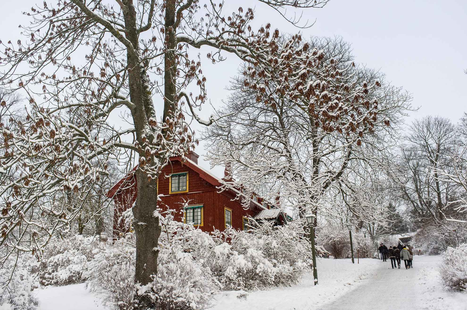Een wit sneeuwlandschap met een rood houten huisje, bomen en enkele wandelaars op de achtergrond.