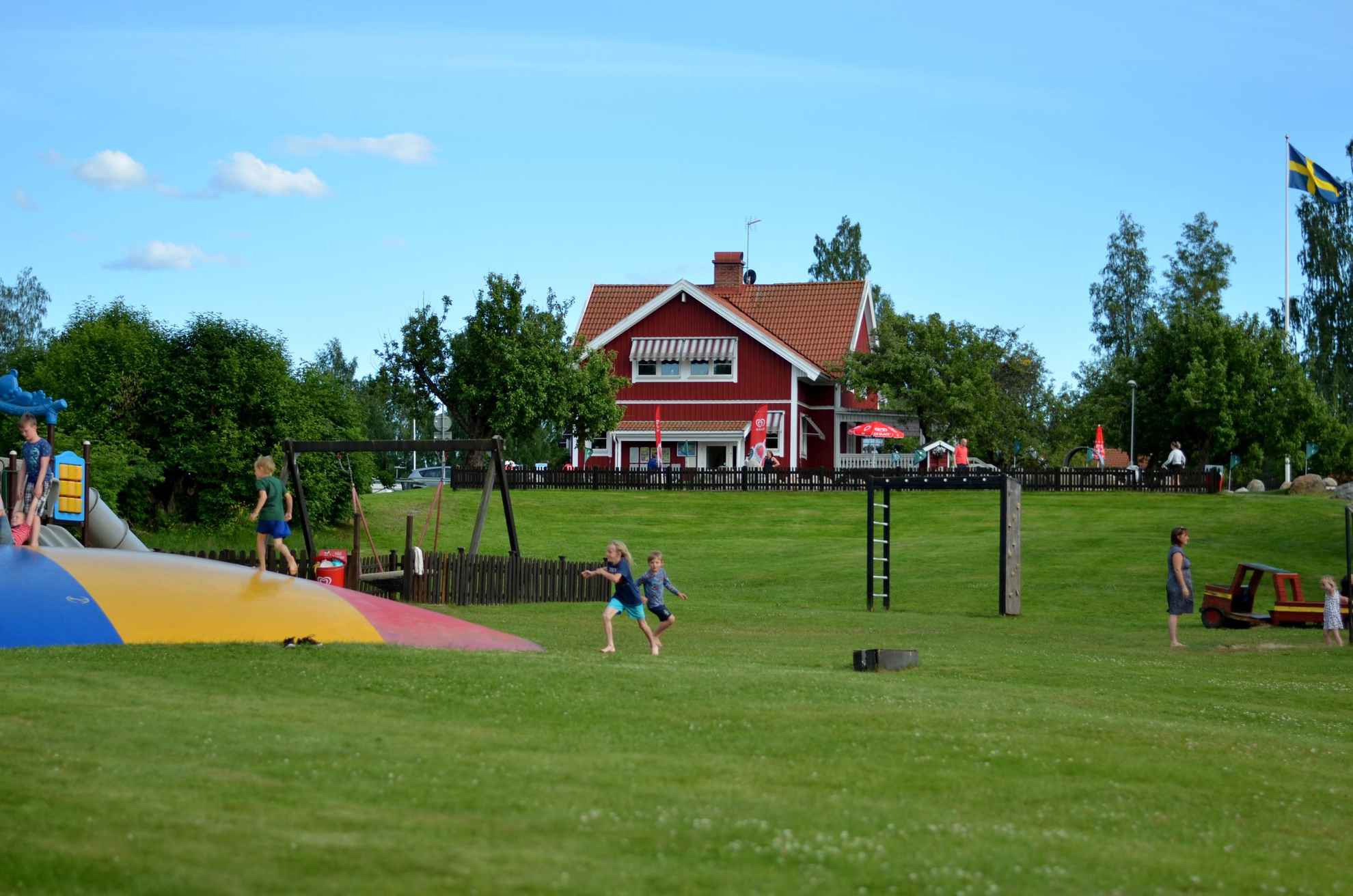 Kinderen op de speelplaats van Frykenbadens Camping. Een groot rood huis achter de speeltuin.