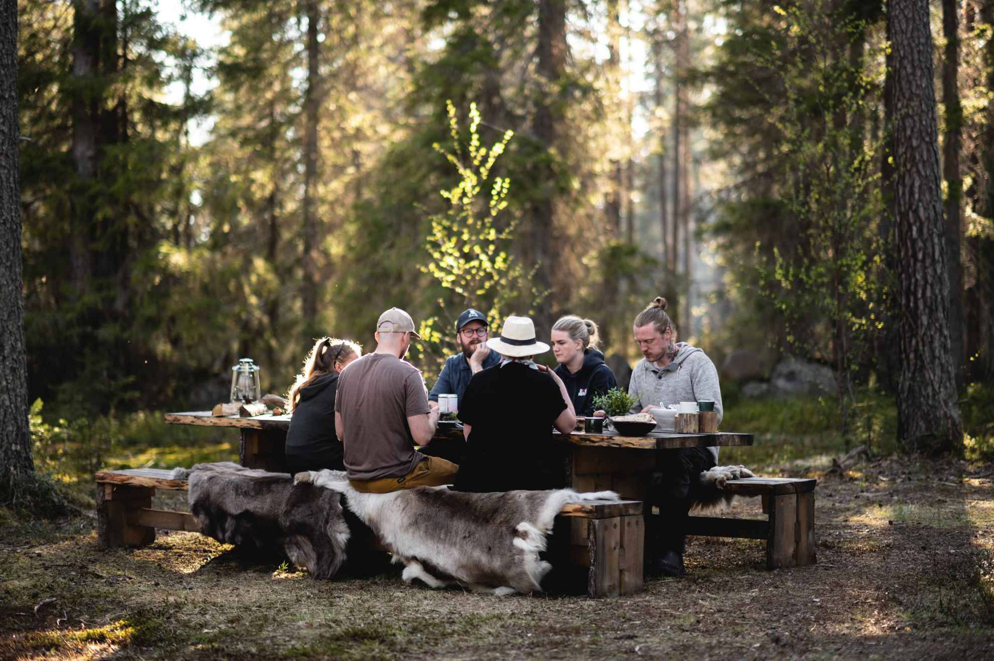 Zes personen zitten aan een houten tafel in een bos. Ze zitten op banken met schapenvachten. De zon schijnt door de bomen.