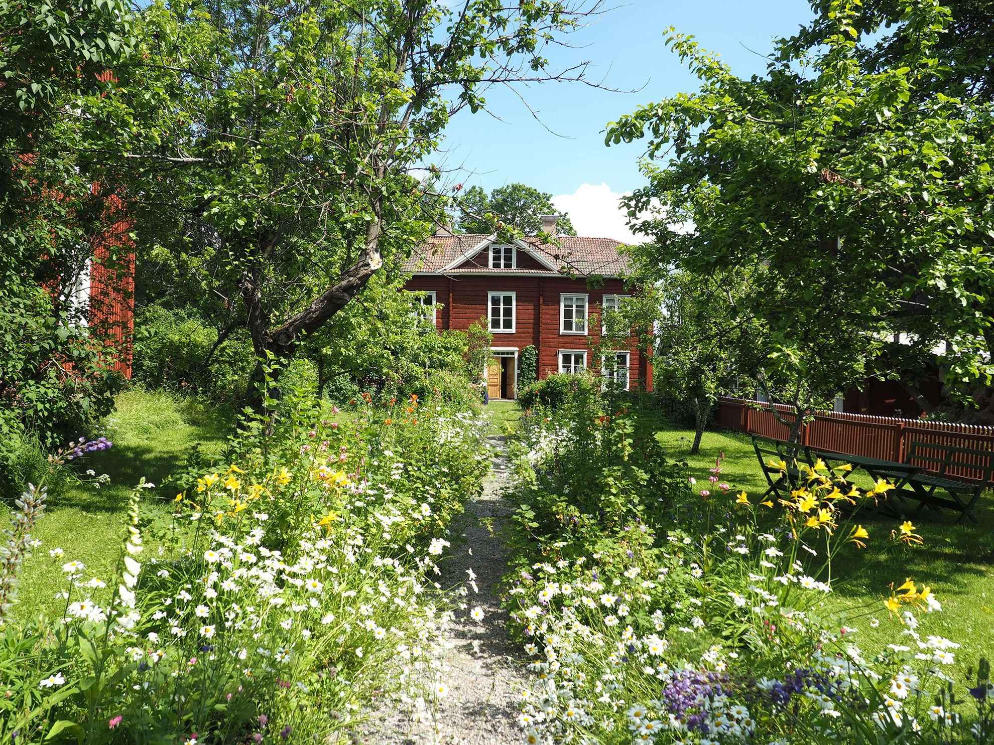 Een onverharde weg met aan weerszijden bloeiende bloemen leidt naar een rood houten huis. Er is tuinmeubilair en bomen op het groene gazon.