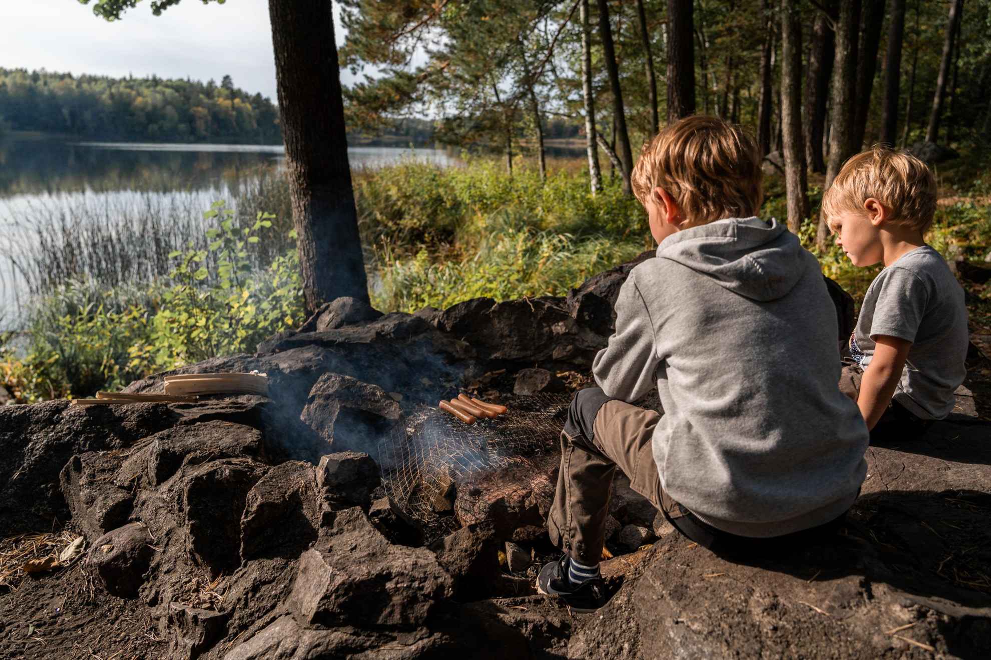 Twee kinderen zitten buiten in de natuur naast een vuurtje worstjes te grillen.