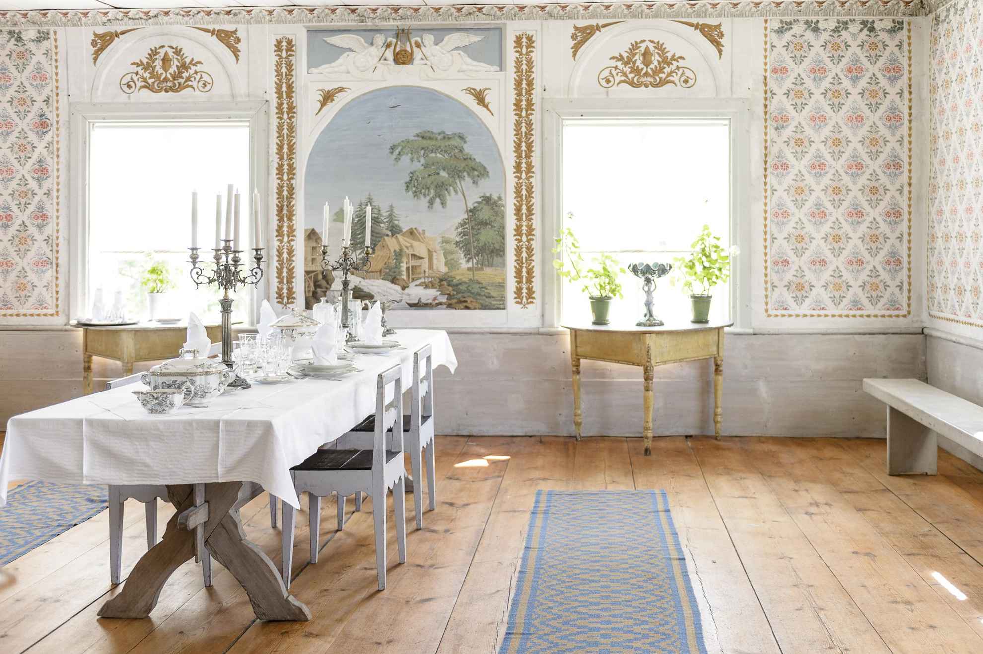 In een oude kamer staat een tafel met bestek, kroonluchters en stoelen. De muren zijn versierd met 19e-eeuwse stencils en daglicht schijnt door de ramen.