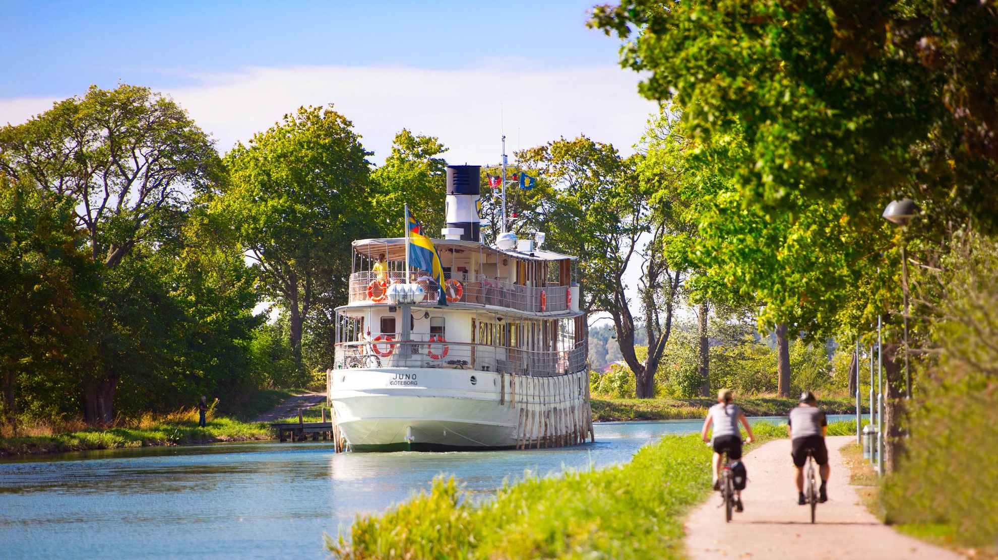 Het is zomer en twee mensen fietsen langs het water van het Götakanaal. Een boot vaart langs het kanaal.