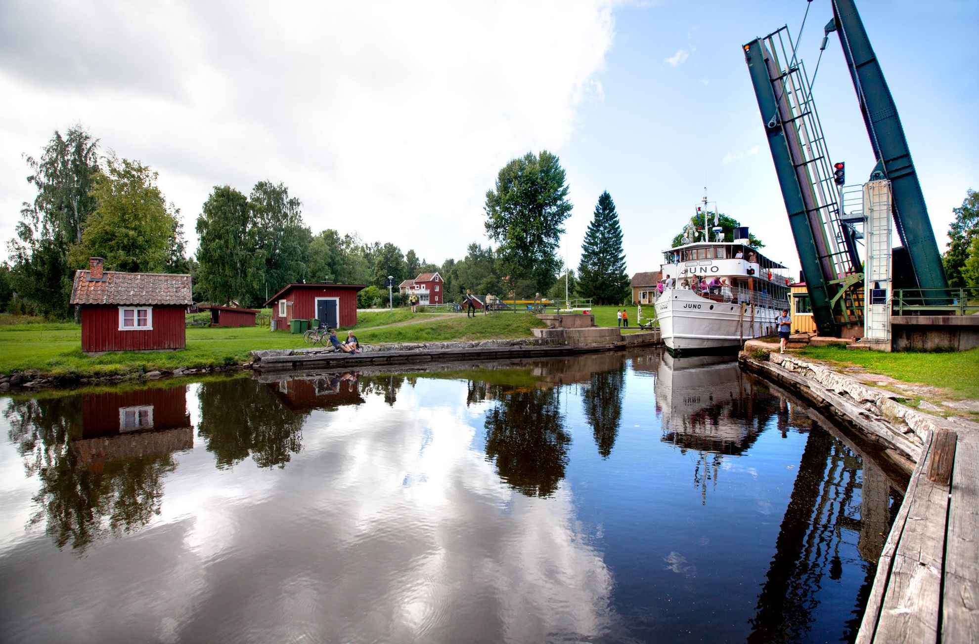 Een grote boot die in de zomer door een sluis in het Götakanaal gaat. Kleine houten huisjes en groen omringen de gracht.