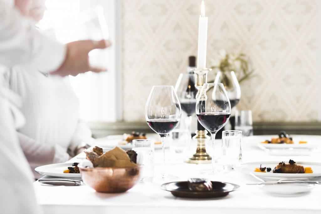 Een gedekte tafel met een wit kleed, een brandende kaars, borden met eten en glazen gevuld met rode wijn. Op de voorgrond van de foto staat een ober.
