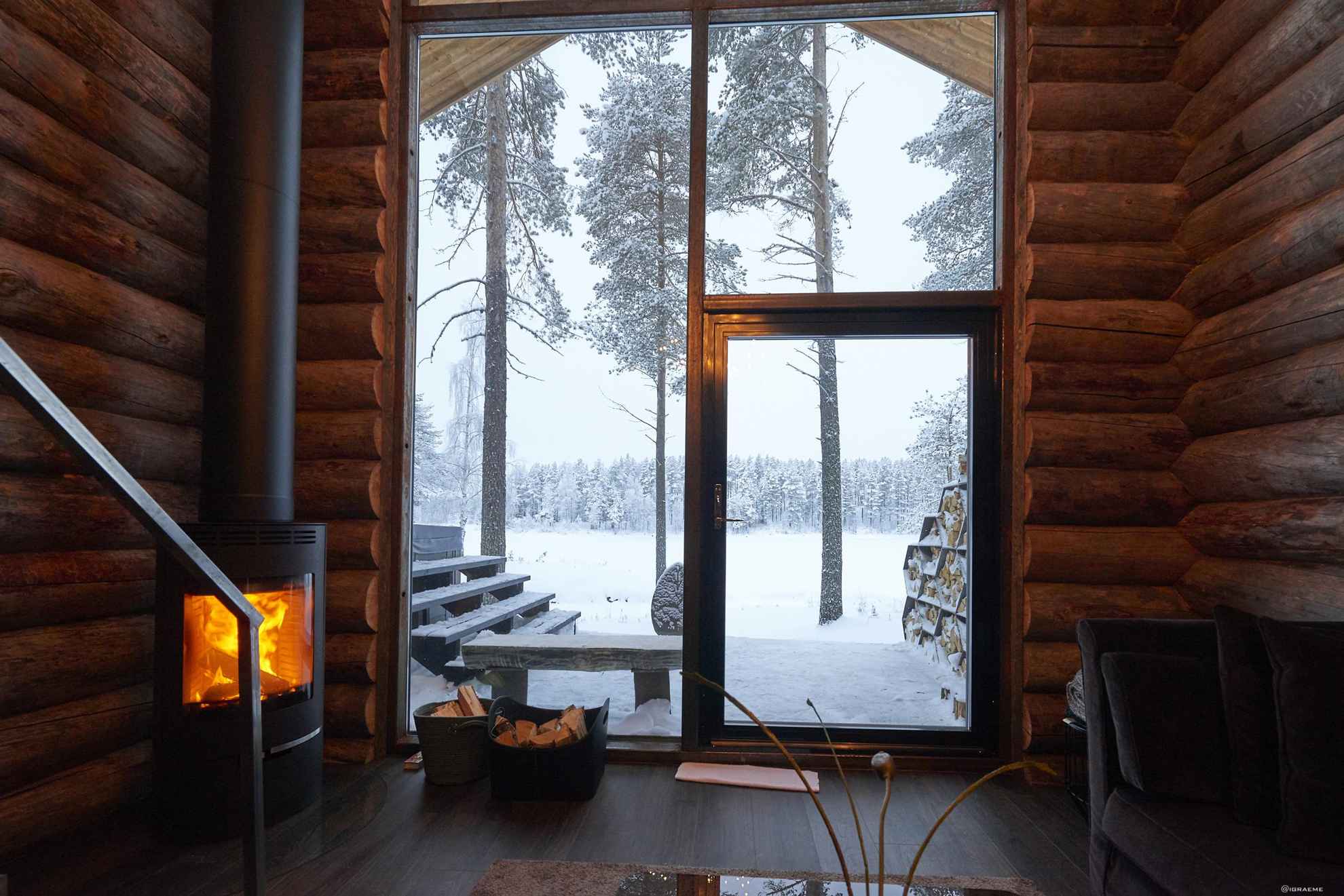 Een houten blokhut met een open haard en grote ramen. Buiten is het winter.