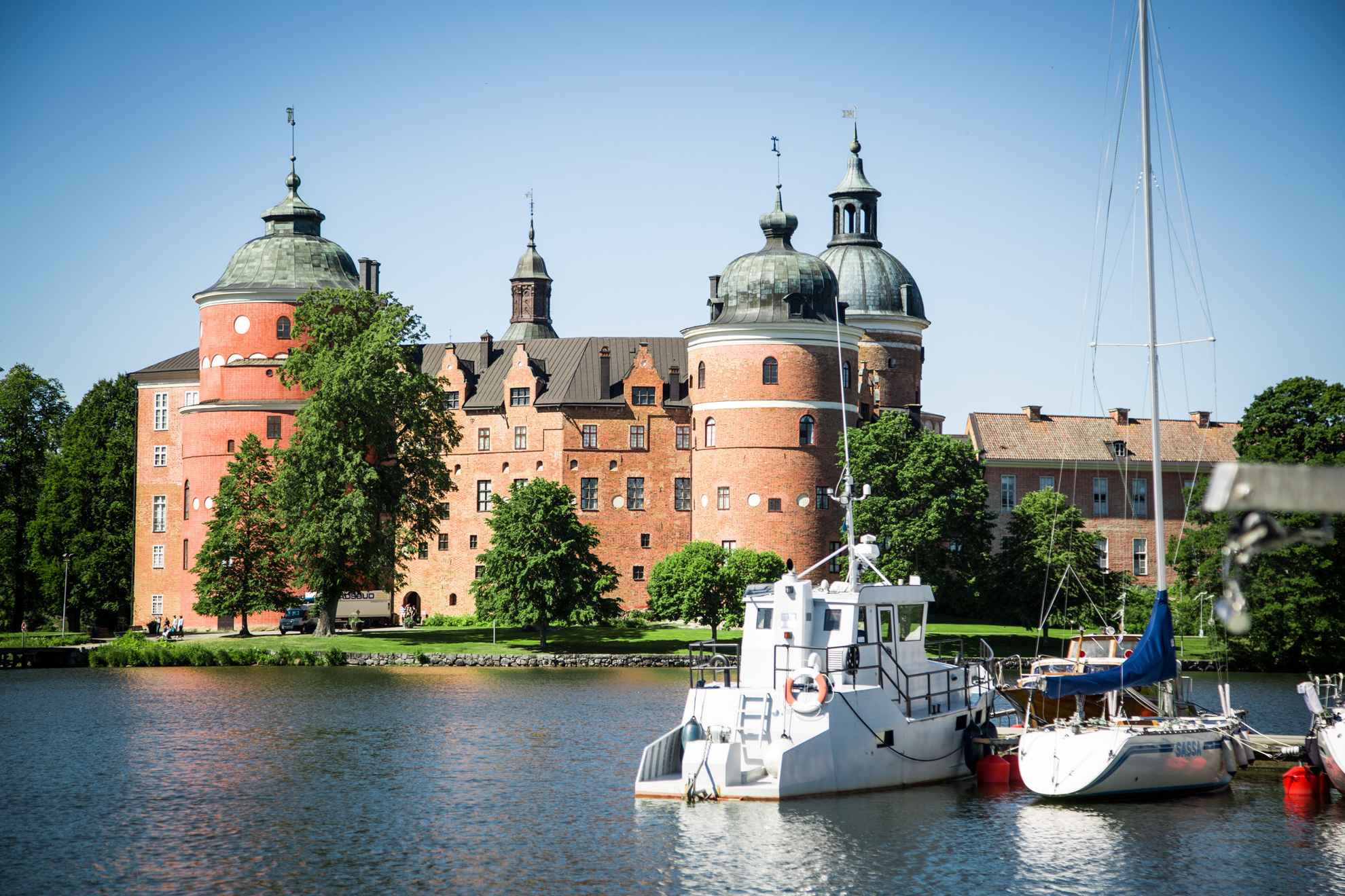 Een kasteel omringd door groen ligt aan de overkant van een meer. De zon schijnt en boten liggen aangemeerd bij de pier.