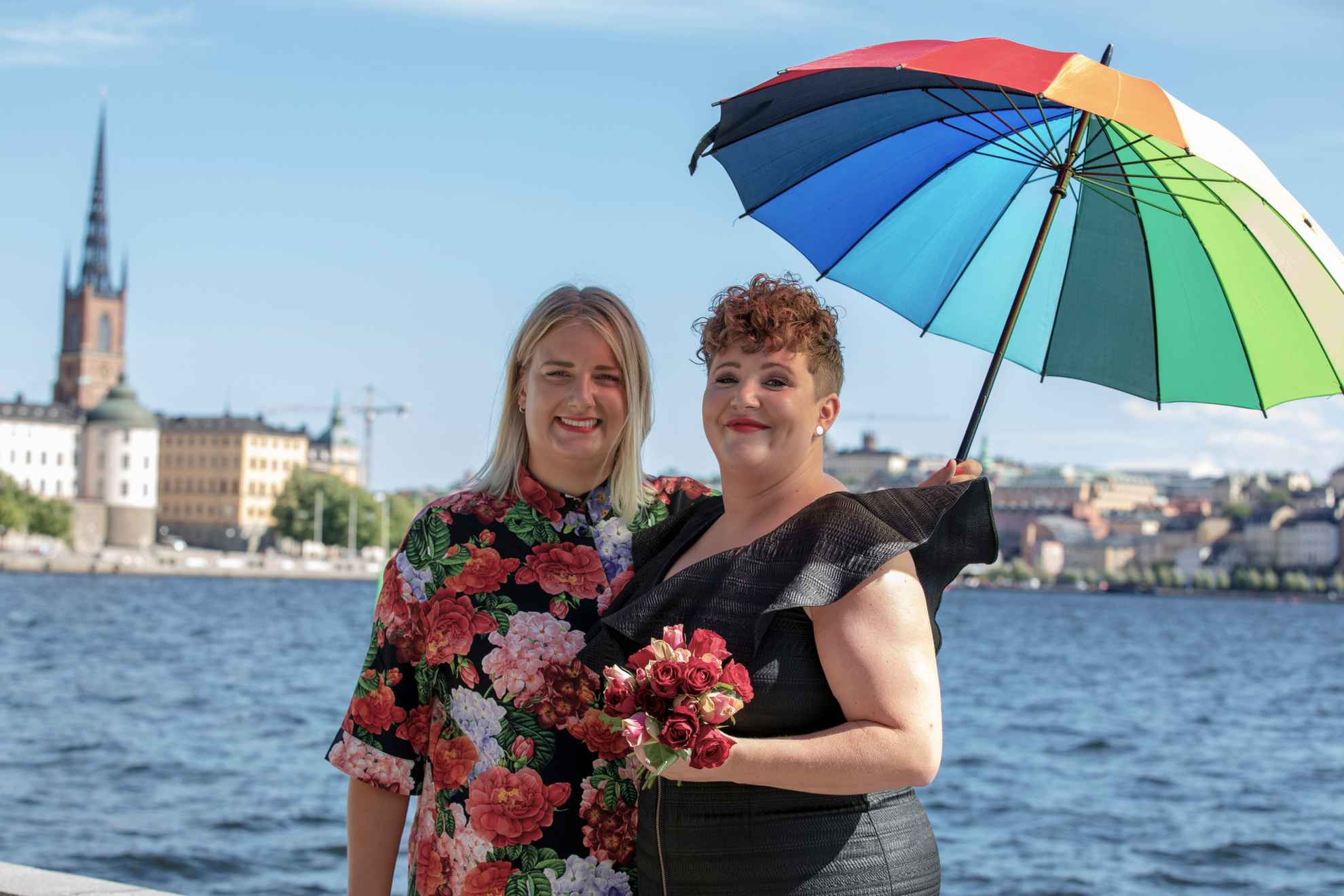 Twee vrouwen, één met een zwarte jurk en één met een shirt met bloemmotief, houden een paraplu vast met de kleuren van de regenboog. Ze staan aan het water met uitzicht op de stad Stockholm op de achtergrond.