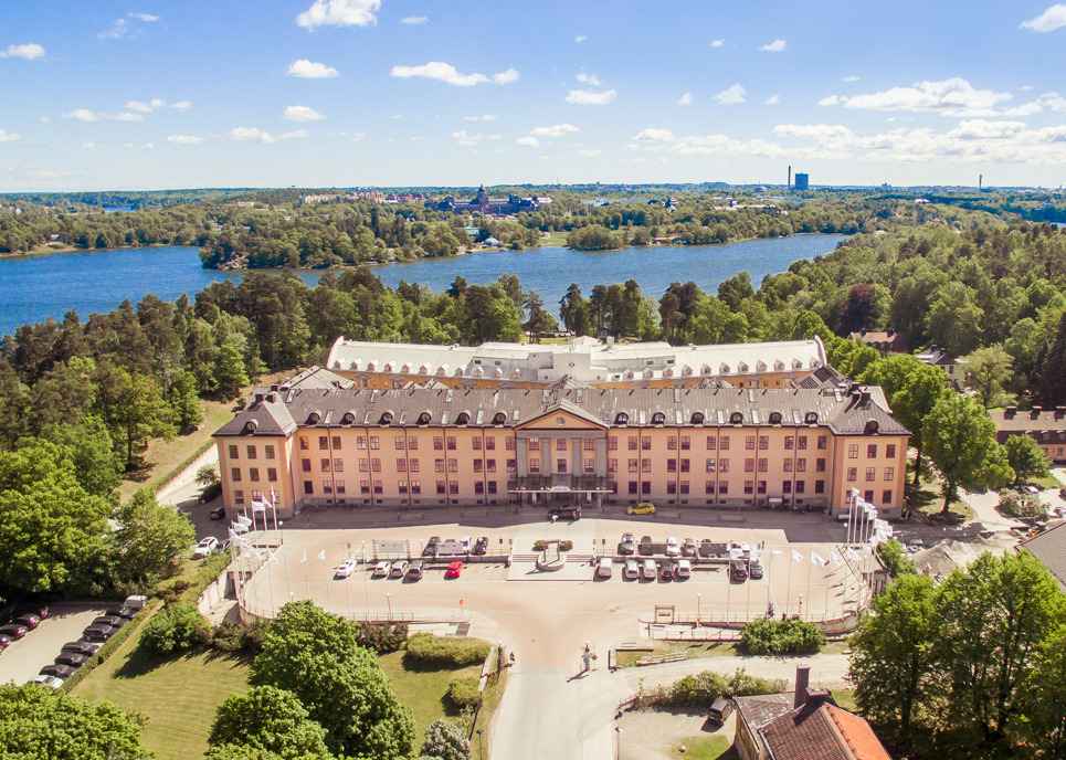 Luchtfoto van Hagastrand met op de achtergrond het glinsterende Brunnsviken en op de voorgrond geparkeerde auto's op een zonnige dag.