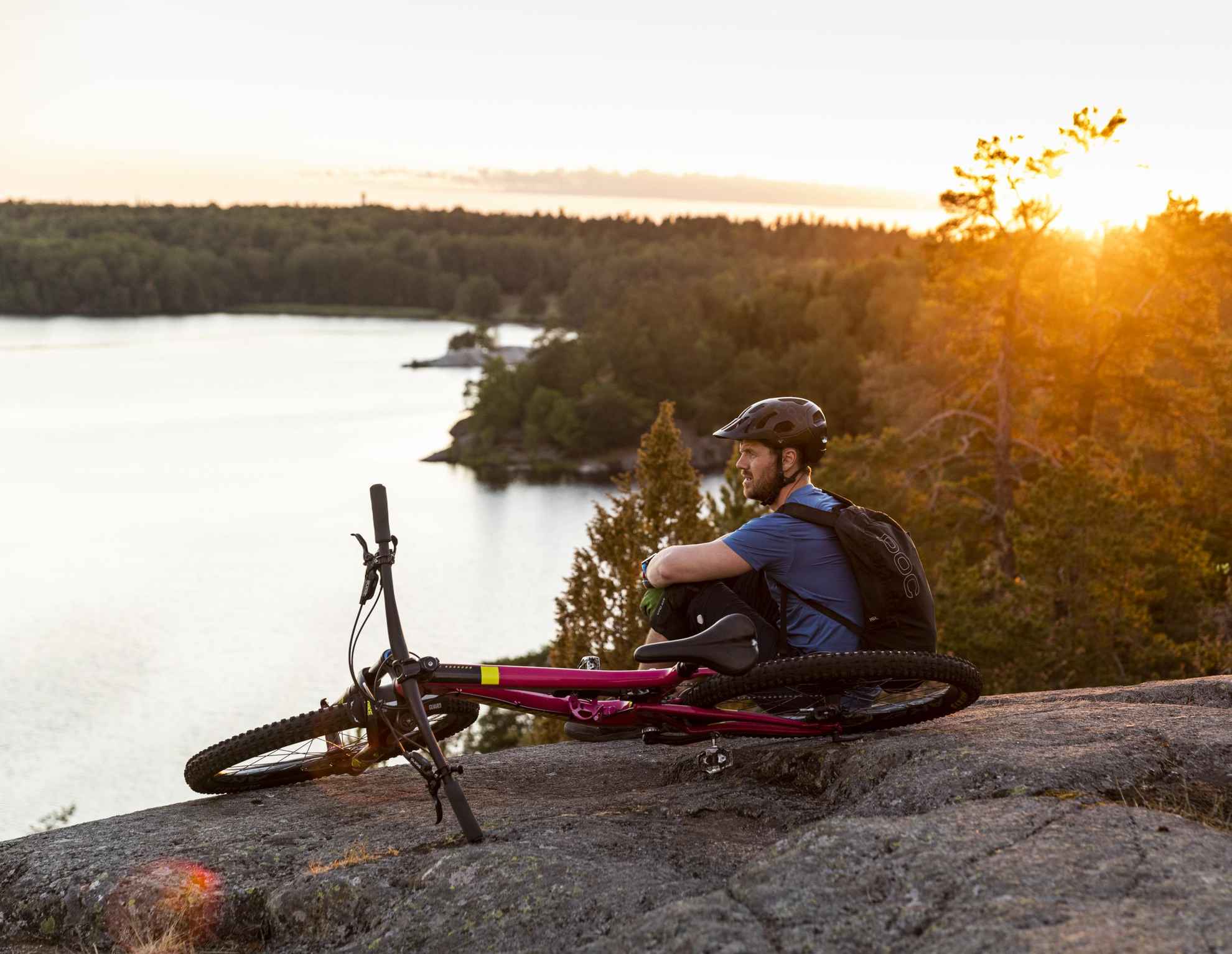 Een man ontspant op een klif met zijn fiets naast hem. De man kijkt uit over een meer.