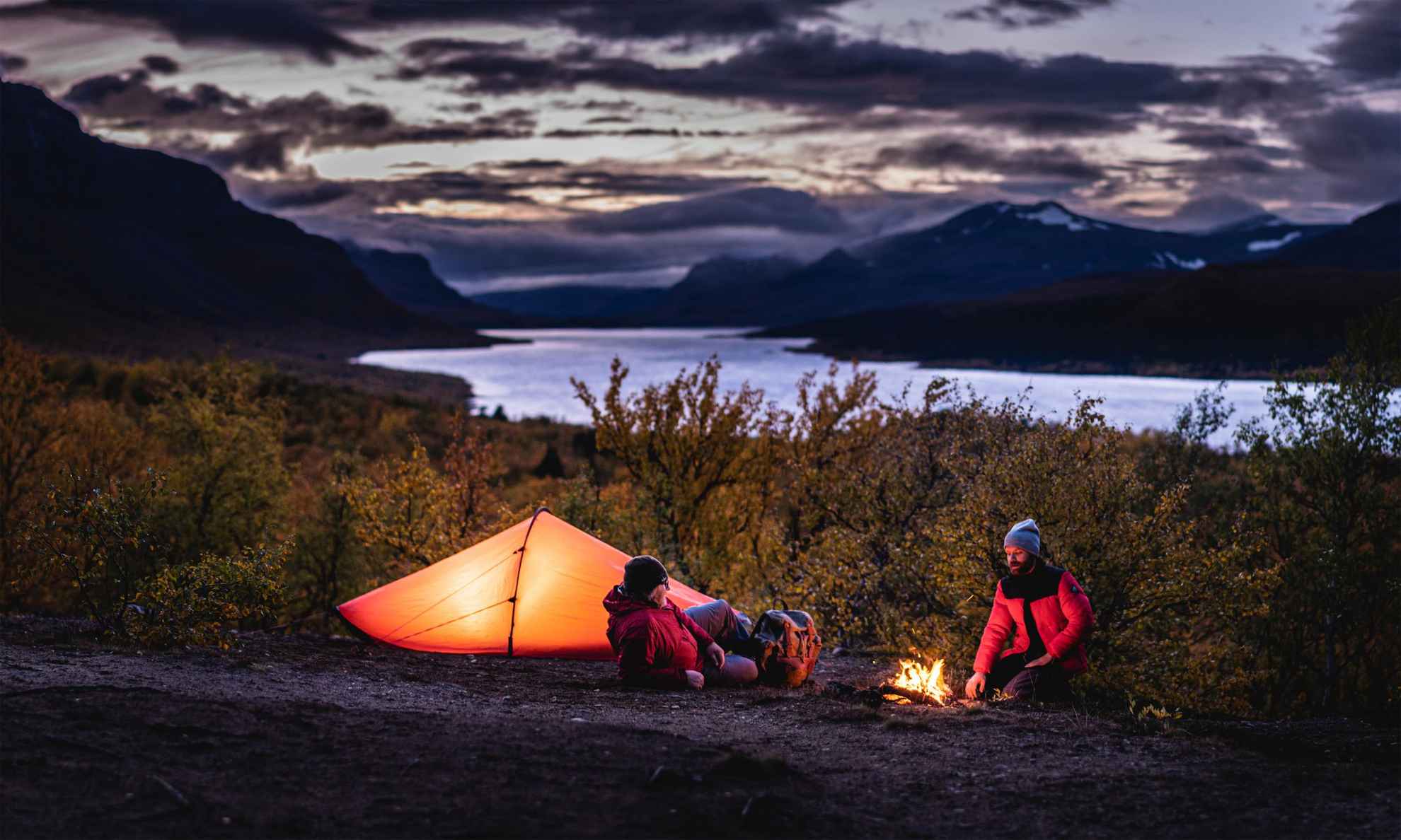 Twee mensen zitten naast een tent en een kampvuur. Een rivier en bergen zijn op de achtergrond.