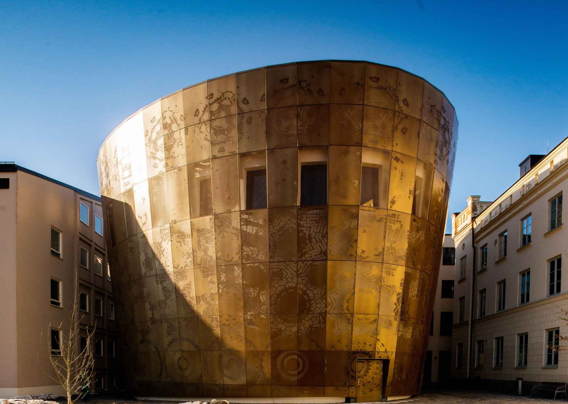 Een subtiel glanzend hoefijzervormig gebouw. In de goud glinsterende gevel zie je een vaag patroon.