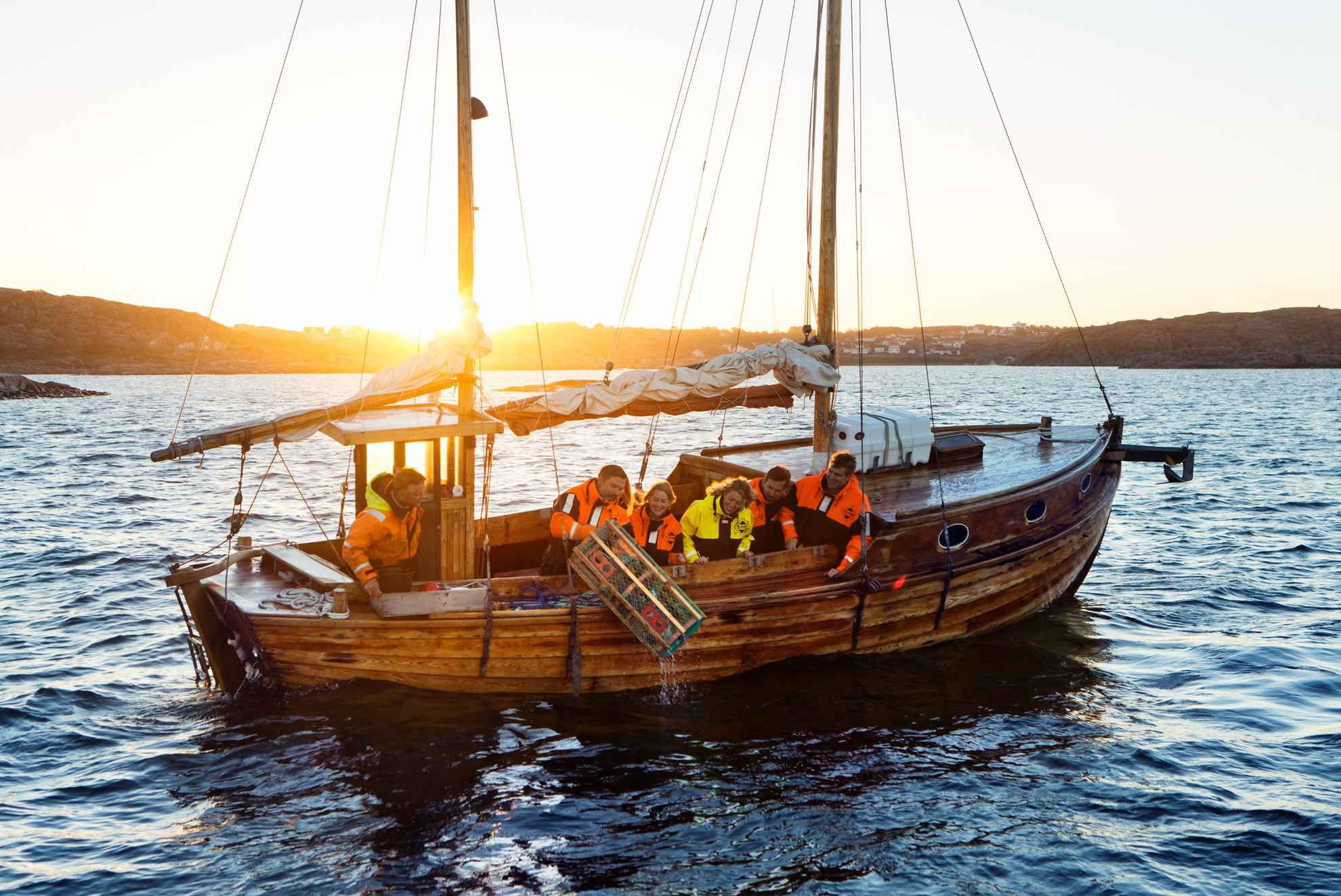 Een houten zeilboot met zes personen, waarvan er één een kreeftenkooi in zee gooit. De zon gaat onder op de achtergrond.