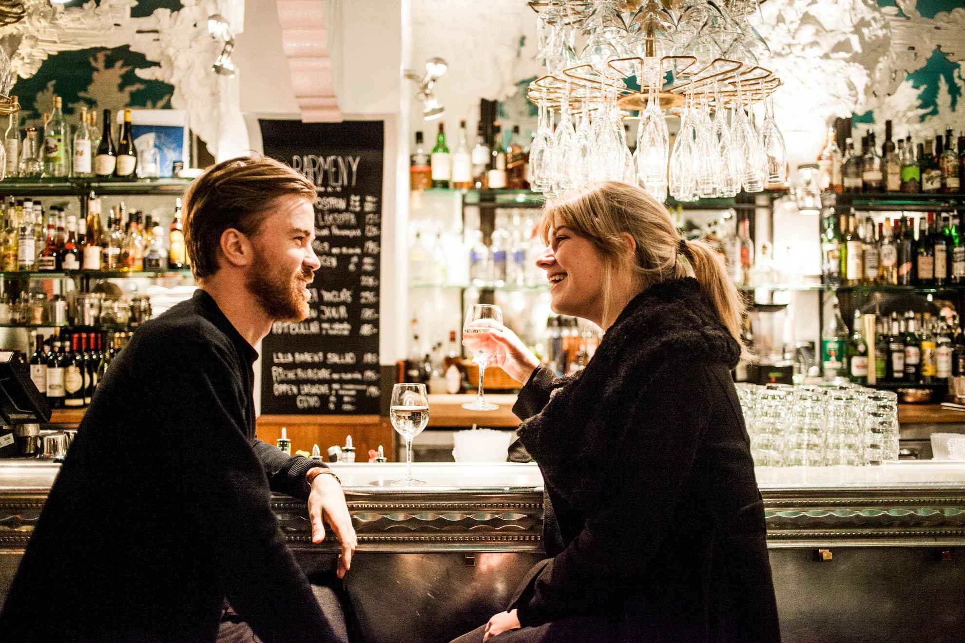 Een man en een vrouw zitten in een goed verlichte bar, elk met een glas wijn.