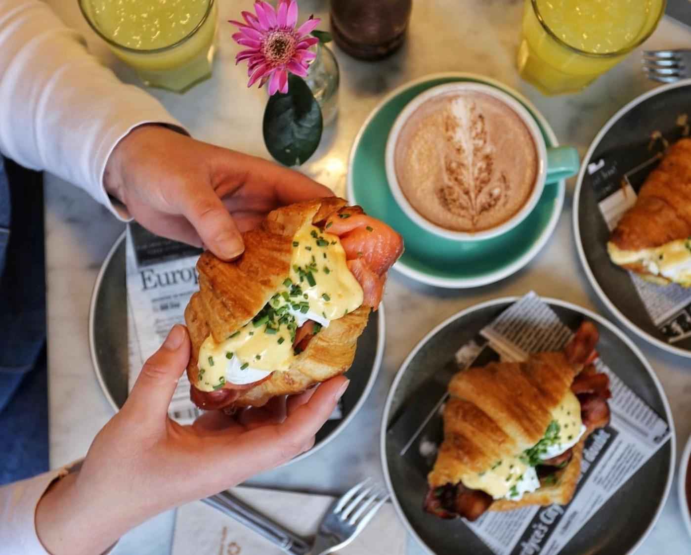 Een persoon houdt een croissant Eggs Benedict vast. Op de tafel staan borden met croissants, glazen met sap en een kopje cappuccino.