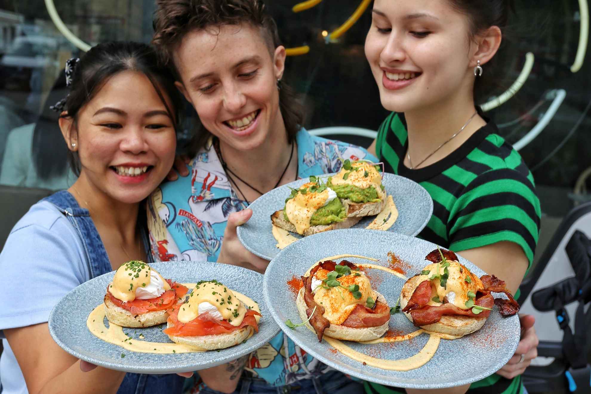 Drie lachende vrouwen houden borden met verschillende sandwiches vast terwijl ze buiten restaurant Greasy Spoon staan.