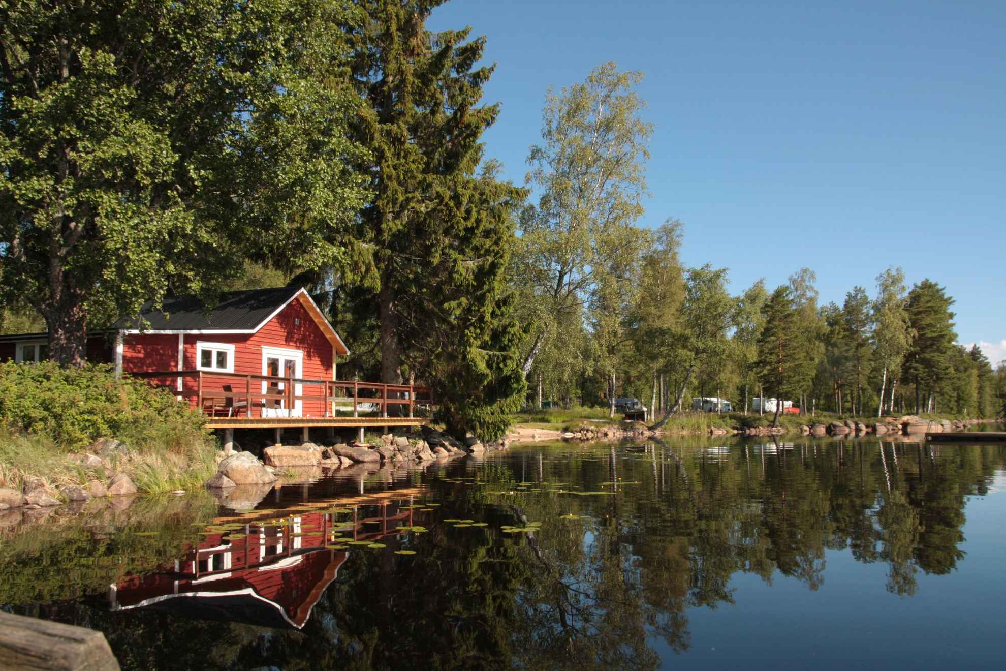 Een rood huisje omgeven door groen, gelegen aan een meer bij Värmlandsgården. Caravans op de achtergrond.