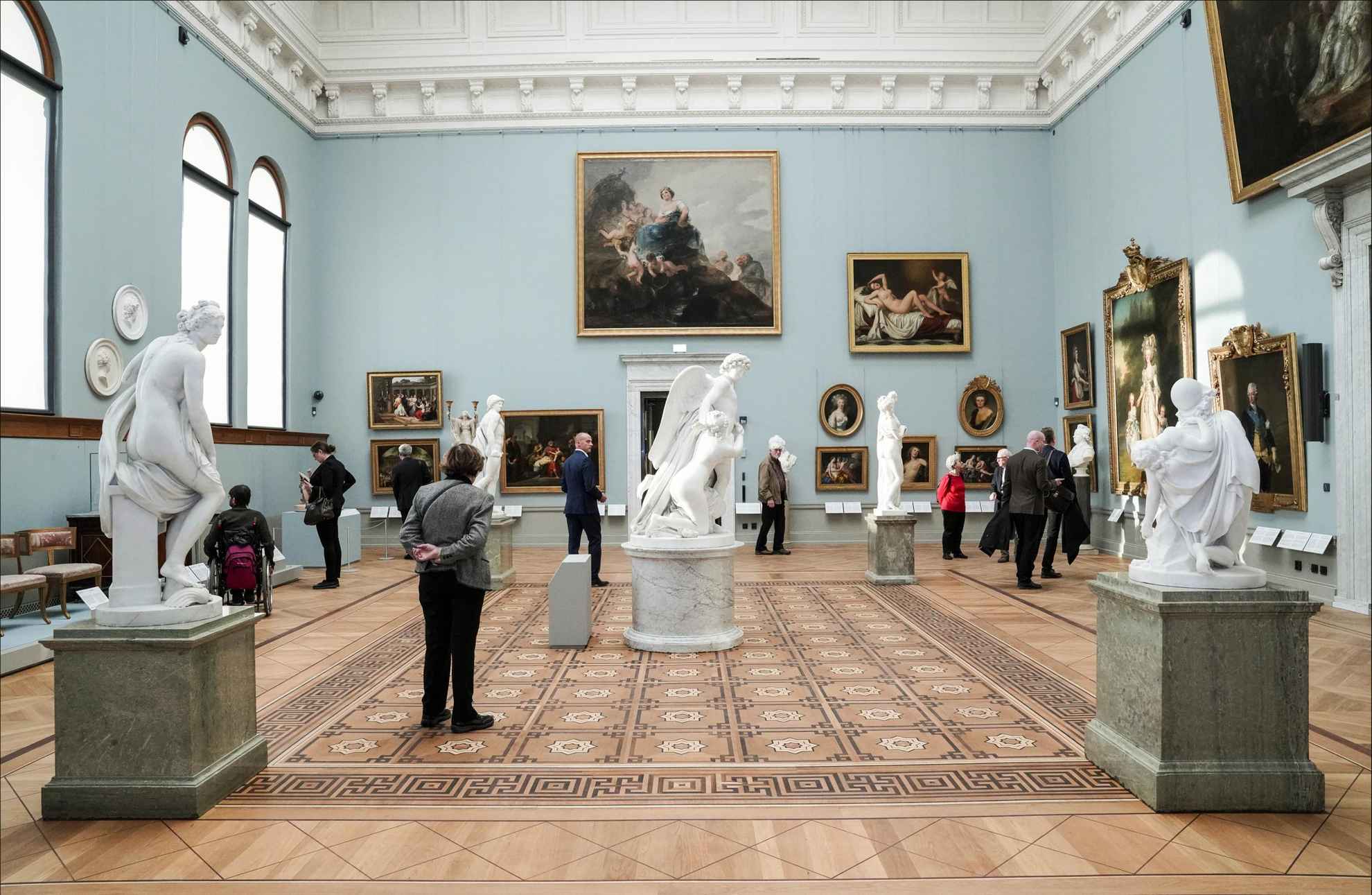 In een grote ruimte met schilderijen en sculpturen lopen mensen rond.