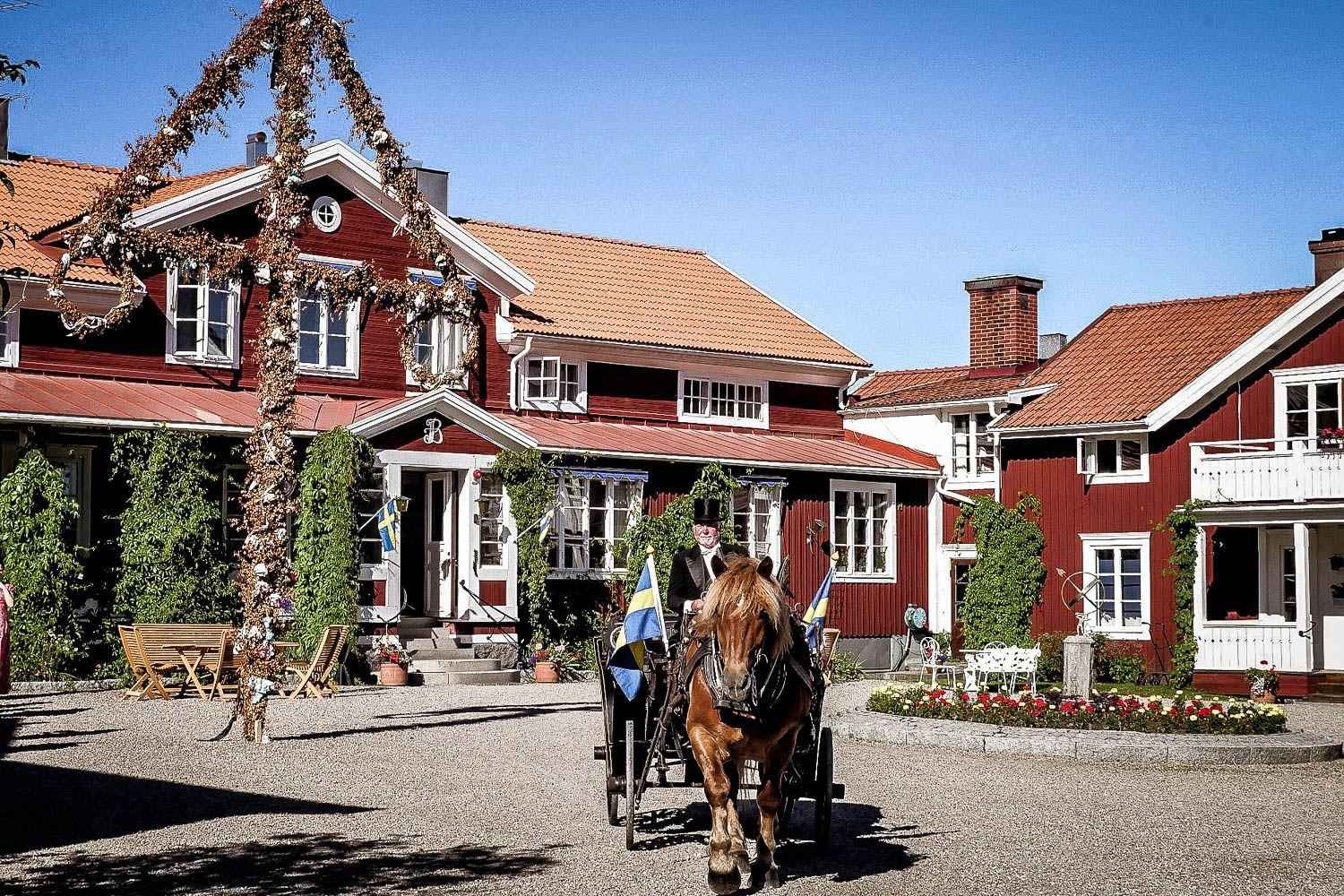 Een groot rood houten huis met witte randen in de zomer. Voor het huis staan een midzomerpaal en een paard met koets met twee Zweedse vlaggen.