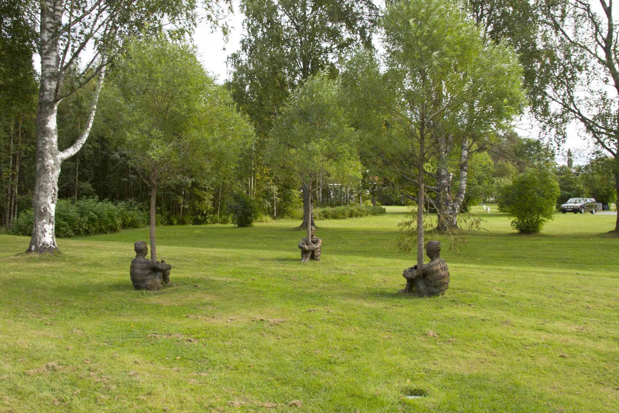 Sculpturen genaamd "Heart of Trees", drie mensen in brons met hun armen en benen gewikkeld rond een boomstam, in een beeldenpark.