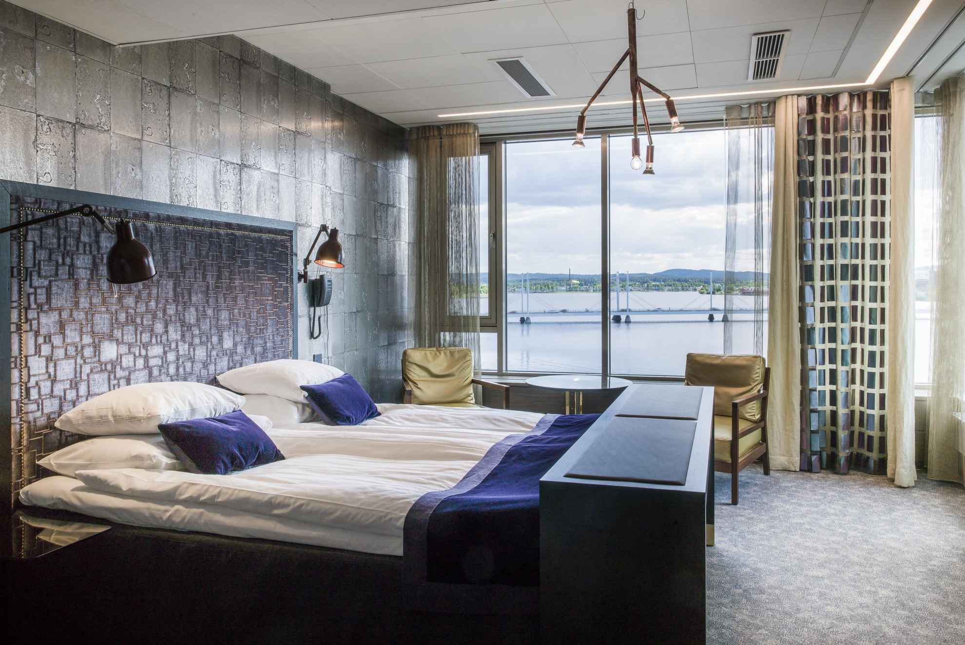 Een hotelkamer met uitzicht op een meer in de zomer. De kamer heeft een tweepersoonsbed, een kleine ronde tafel en twee stoelen.