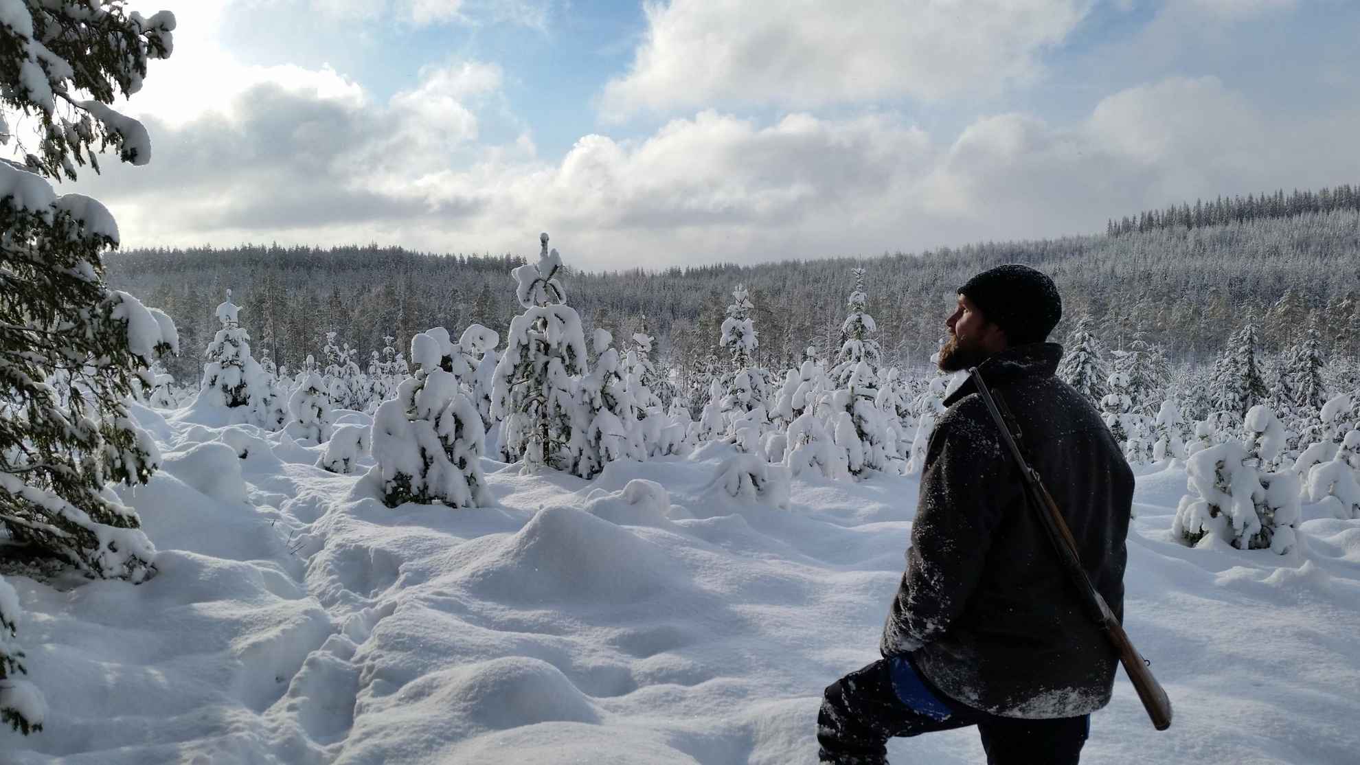 Een man staat in een besneeuwd bos met een jachtgeweer op zijn rug.