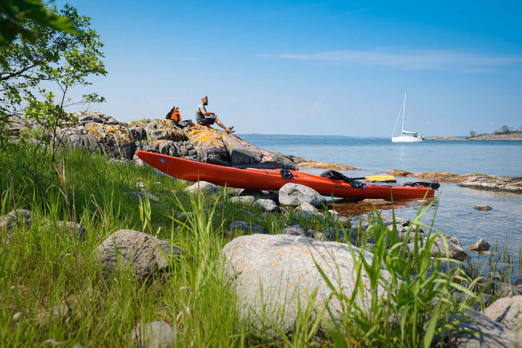 Een oranje kajak ligt aan de kant van het water terwijl een man geniet van een lunch op de kliffen. Op de achtergrond ligt een witte zeilboot.