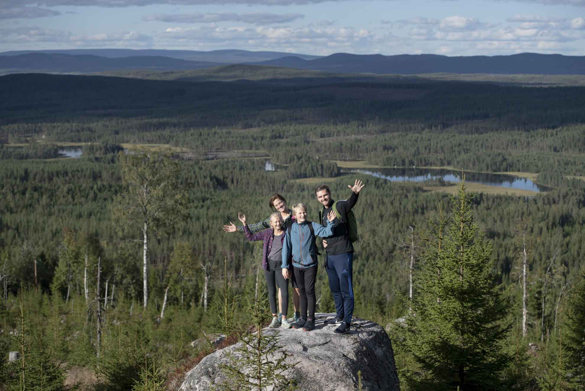 Een gezin van vier staande op een rots, zwaaiend naar de camera. Bos, meren en bergen achter hen.