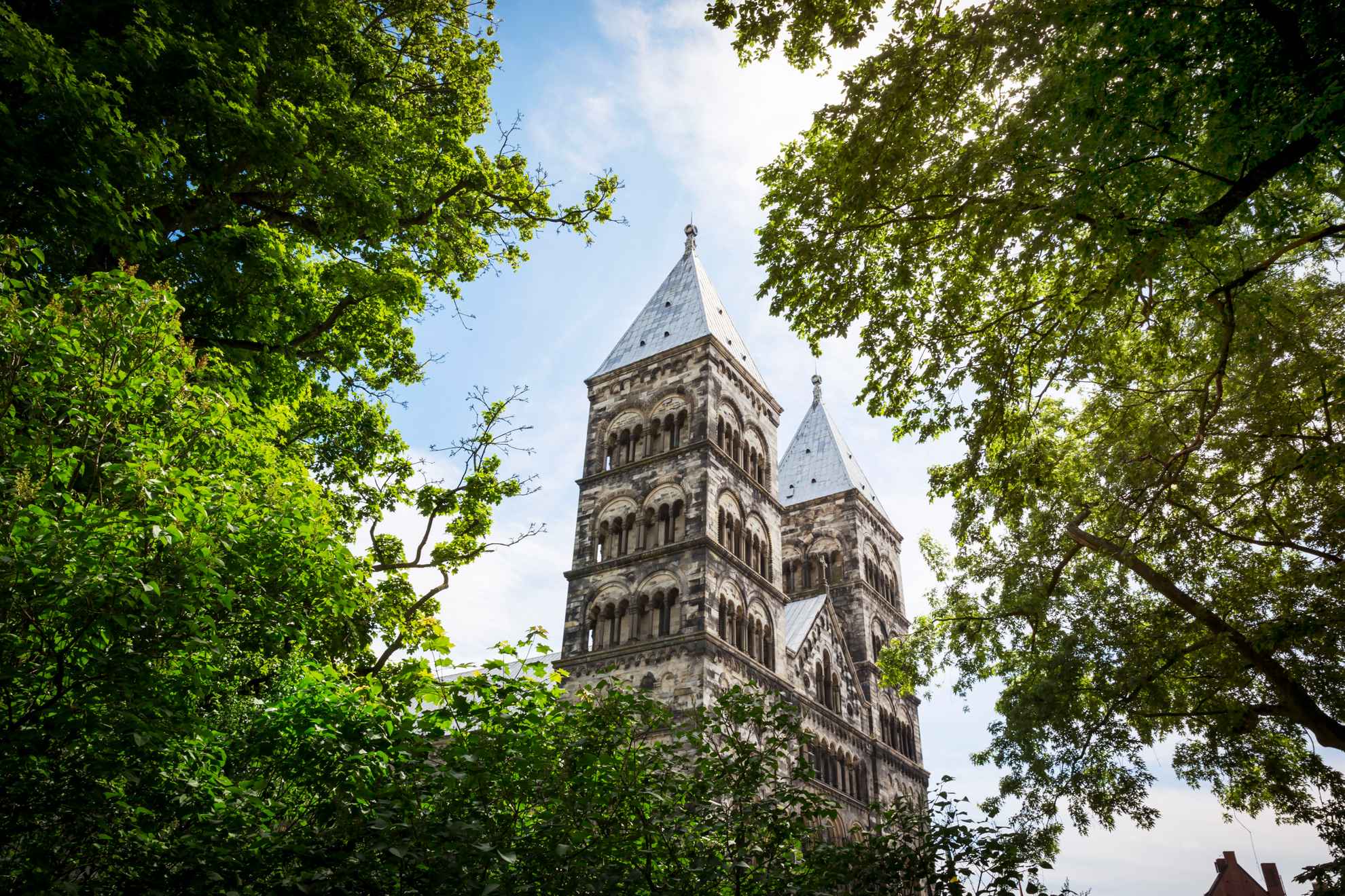 Een foto in kikvorsperspectief van de torens van de stenen kathedraal van Lund, genomen van tussen de bomen.