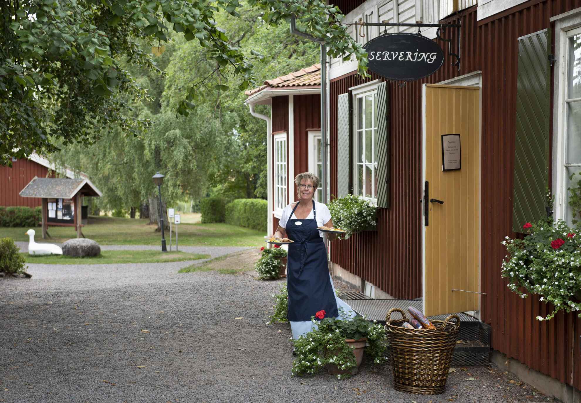 Een vrouw in een blauw schort met een bord in elke hand, staat bij de ingang van een rood houten huis.