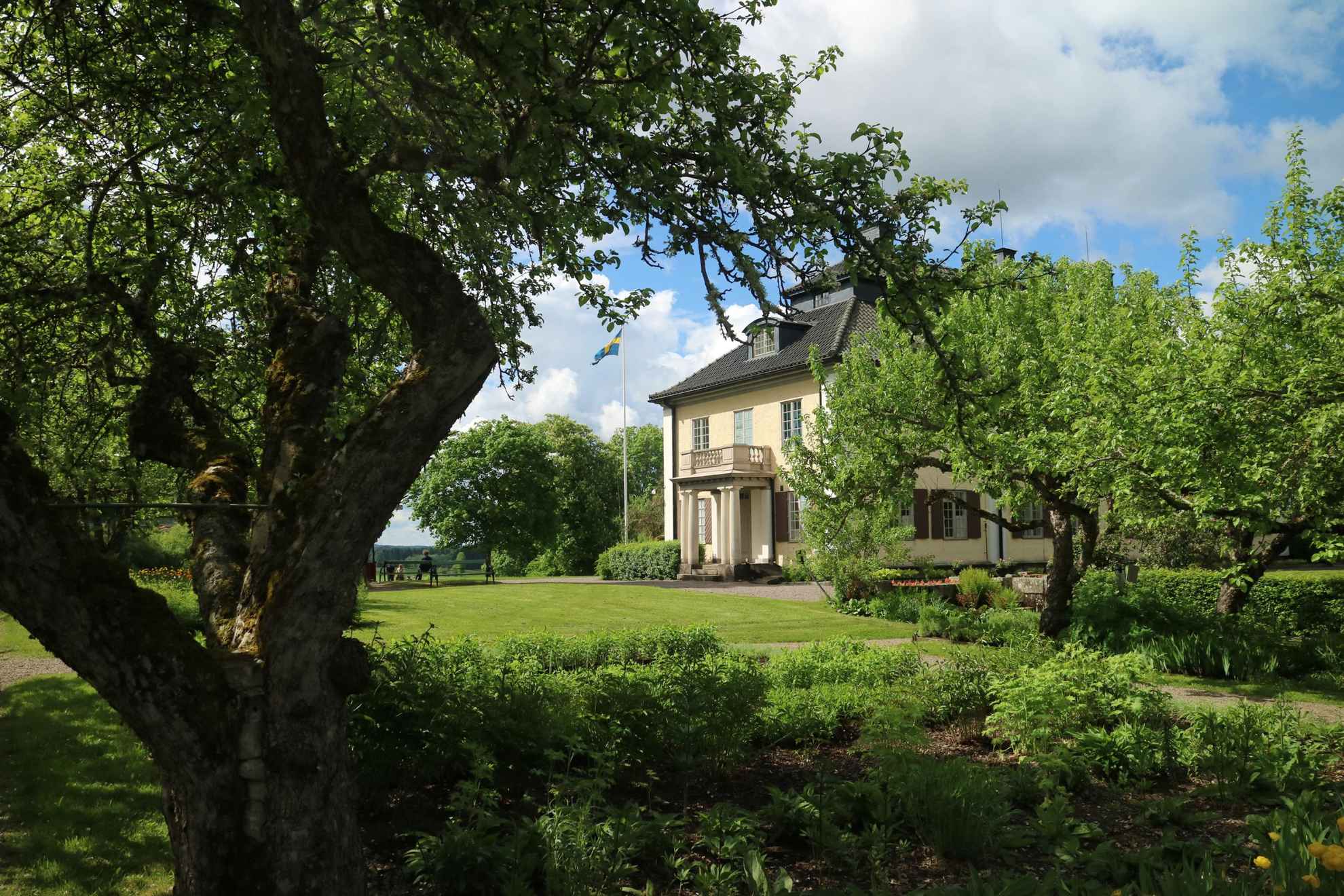Een mooi lichtgeel stenen huis met een tuin. Voor het huis een groen gazon en rondom het huis staan bomen, struiken en bloemperken.
