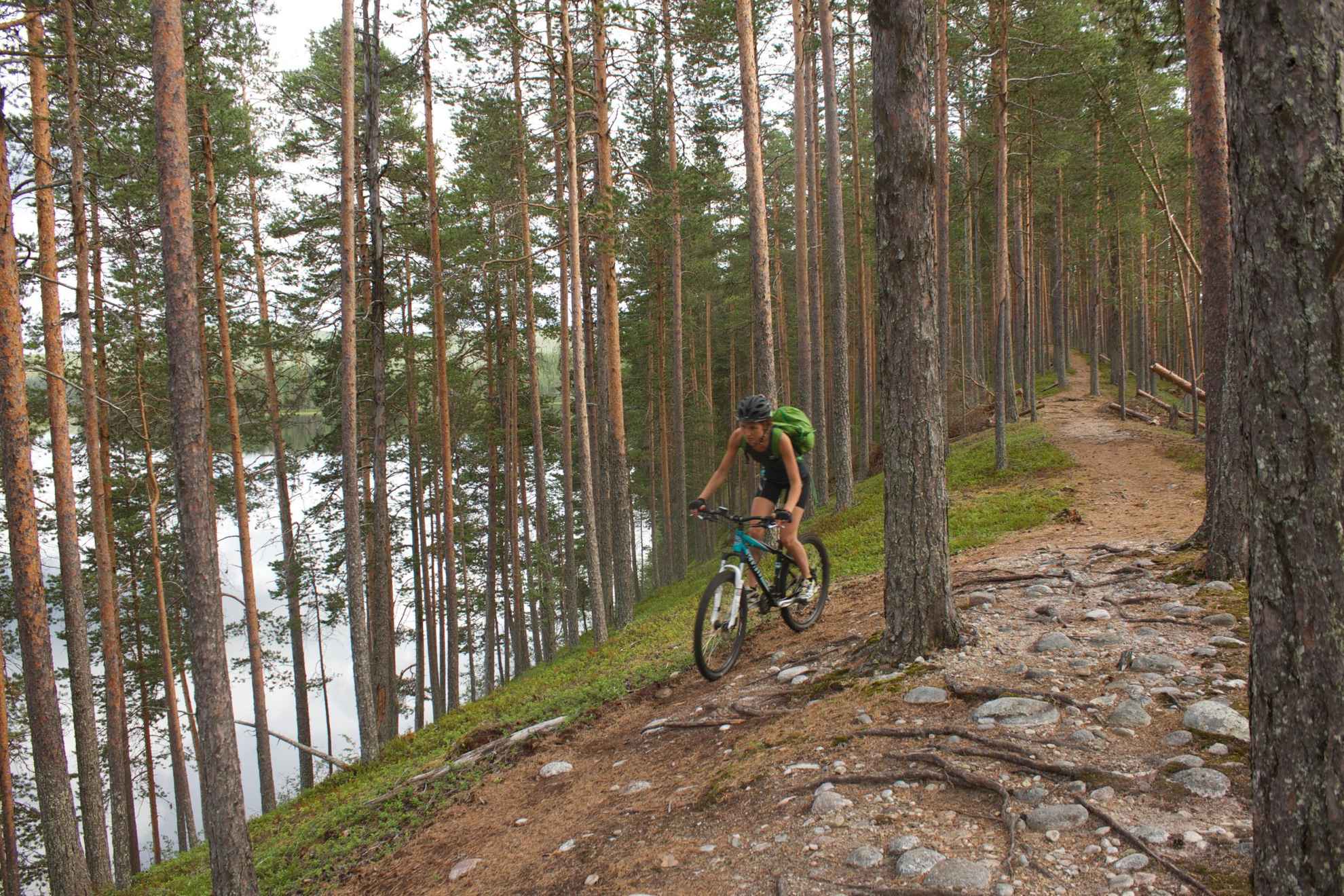 Een vrouw met een rugzak fietst in een dennenbos. Je ziet een meer tussen de bomen.