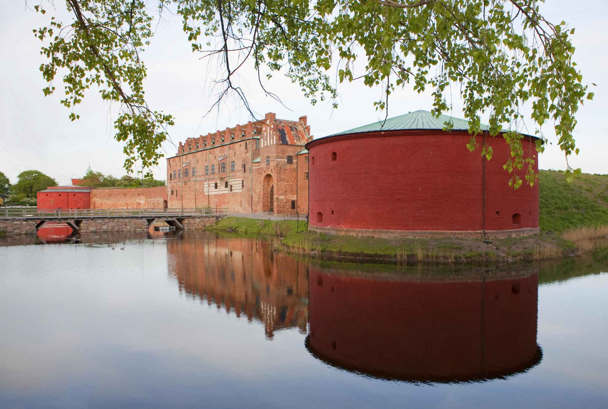 Kasteel Malmöhus is een grote ronde toren naast een bakstenen gebouw, omgeven door water. Een andere ronde toren aan de andere kant op de achtergrond. Groene bladeren op de voorgrond.